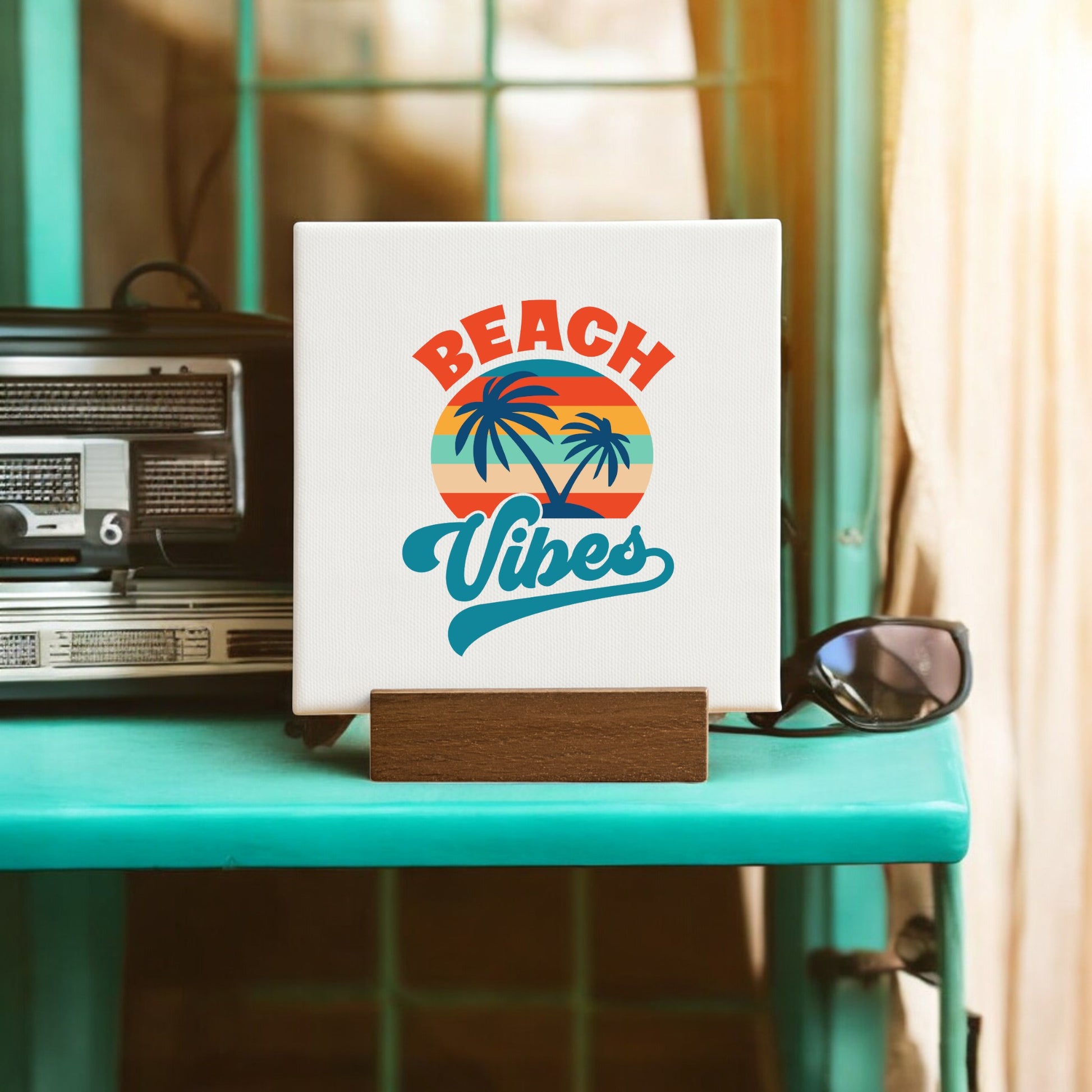 The Mirabilia Boutique Beach Vibes Sunset Canvas – Bold Tropical Mini Sign displays a sunset, palm trees, and "Beach Vibes," standing on a turquoise shelf by a vintage radio and sunglasses with sunlight through beige-curtained windows.