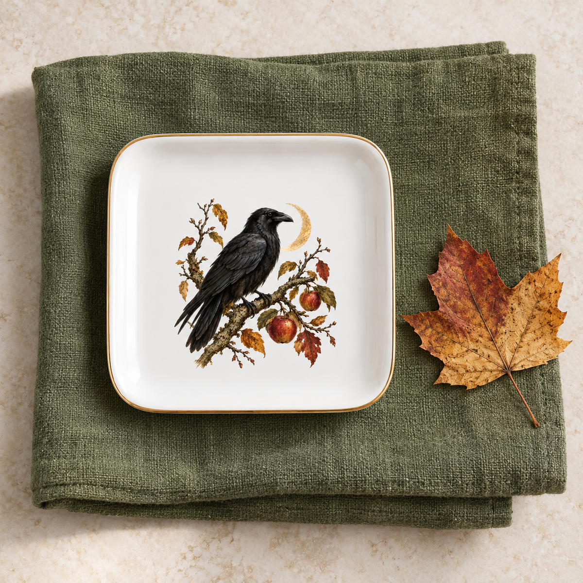The Orchard Sentinel Raven Jewelry Tray by Mirabilia Boutique features a raven with apples and leaves on white ceramic, shown atop a green napkin with a dried maple leaf nearby—ideal for orchard decor or jewelry display.