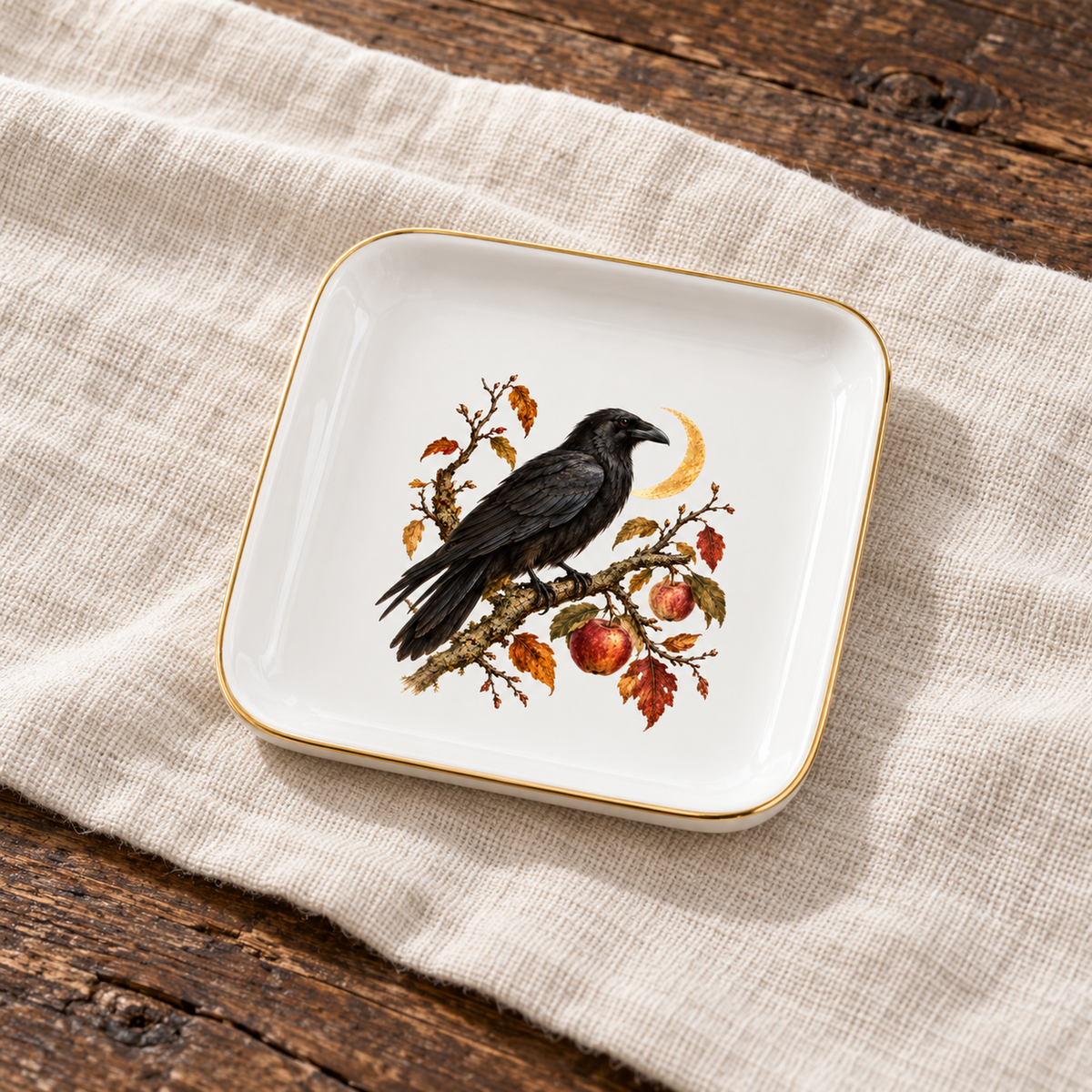 The Orchard Sentinel Raven Jewelry Tray by Mirabilia Boutique features a black bird amid red apples and leaves, gold trim, and rests elegantly on a beige cloth over rustic wood.