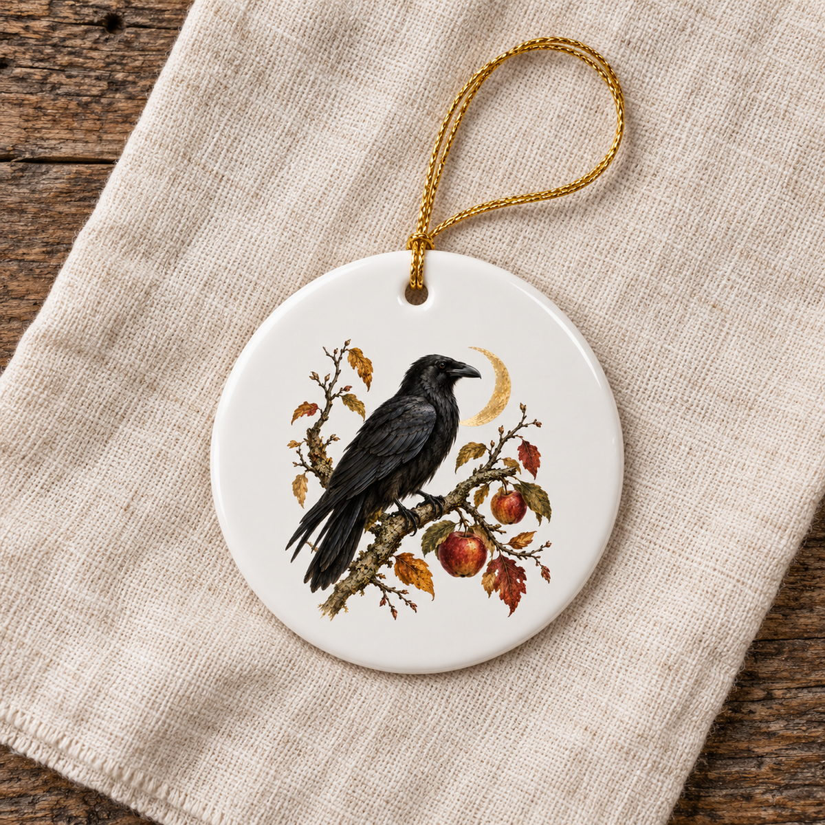 The Orchard Sentinel Raven Ceramic Ornament by Mirabilia Boutique features a black raven among autumn leaves, red apples, and a golden crescent moon on round white ceramic with a gold hanging string.