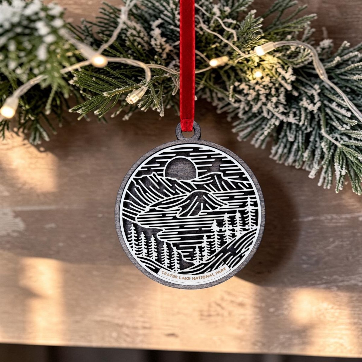 The Crater Lake National Park Christmas Ornament by Mirabilia Boutique features black and white landscape line art on an oval wooden ornament, hanging from a red ribbon in front of snowy pine branches and glowing string lights. The ornament is inscribed with “Crater Lake National Park.”.