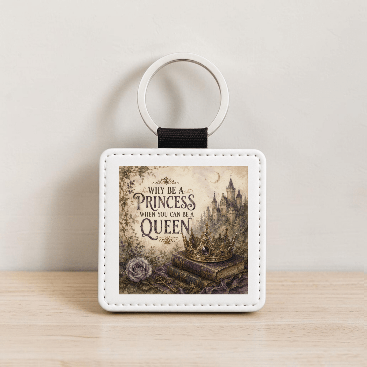 Introducing the "Why Be a Princess When You Can Be a Queen Keychain" by Mirabilia Boutique—a square PU leather keychain with elegant white stitching and a silver ring. This whimsical accessory features an enchanting illustration of a castle, crown, rose, and old books alongside the inspiring queen quote: "Why be a princess when you can be a queen.