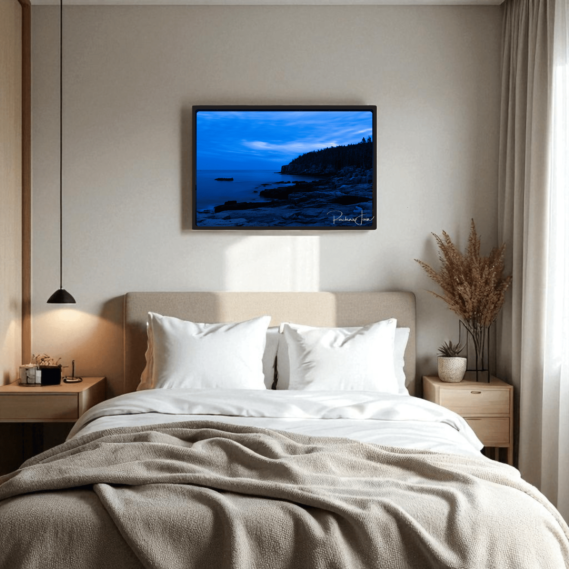 A cozy bedroom features a beige bed with white pillows and blanket. Above, the Otter Point Acadia | Blue Hour Framed Art Print by Mirabilia Boutique adds serene coastal style, complemented by nightstands, pendant light, and coastal wall art.
