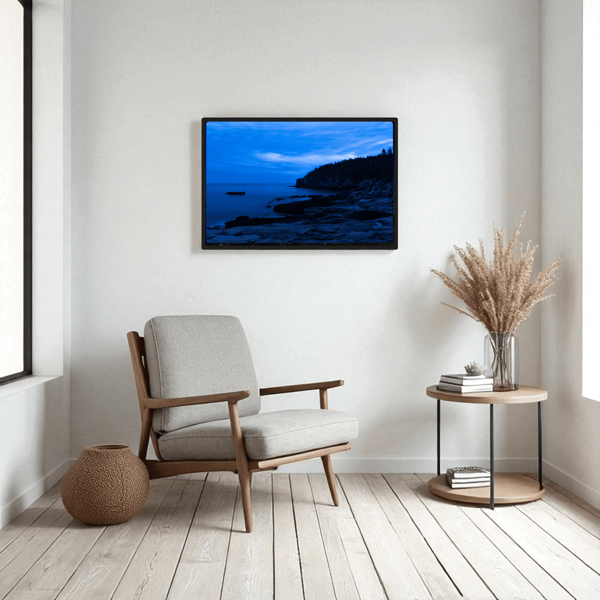 A modern, minimalist living room features a light gray armchair, round side table with a vase of dried grass, wicker basket, and Mirabilia Boutique’s "Otter Point Acadia | Blue Hour" coastal framed art print above the chair. Light wood floors and neutral decor complete the look.