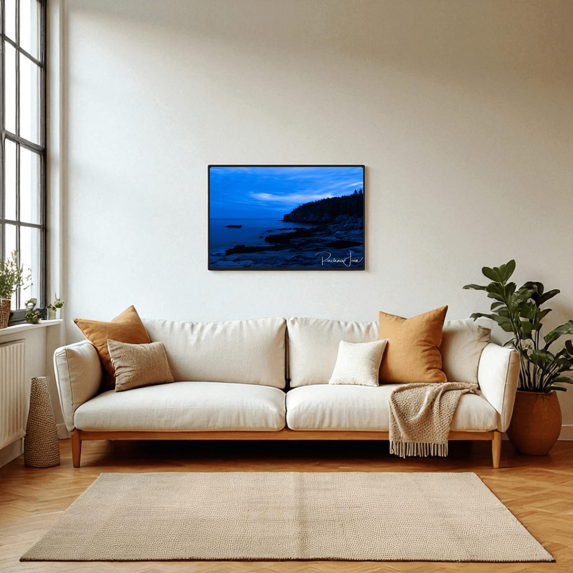 A bright living room with a white sofa, tan and brown cushions, beige throw, and potted plant. Above the sofa hangs Mirabilia Boutique's "Otter Point Acadia | Blue Hour" coastal art print. Large windows fill the space with natural light.