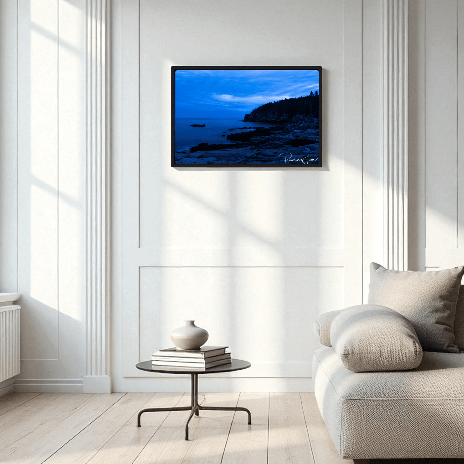 A modern living room with white paneled walls, a light gray sofa, and a round black coffee table displays Mirabilia Boutique’s Otter Point Acadia | Blue Hour Framed Art Print—coastal photography as striking wall art.