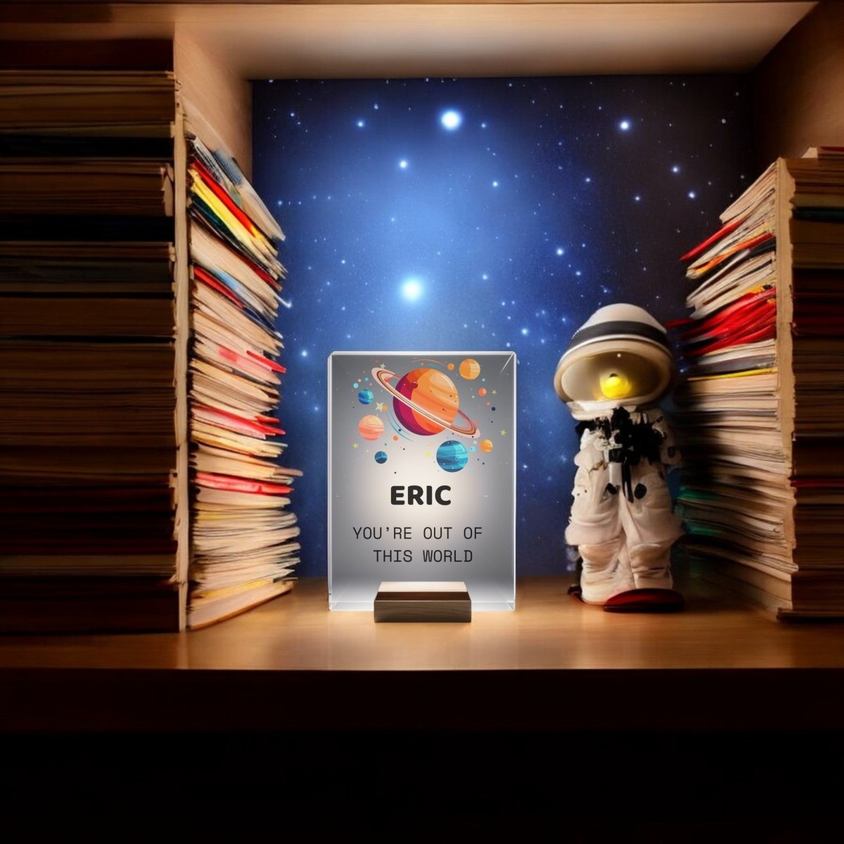 A desk shelf holds stacked books, a small astronaut figurine, and the Mirabilia Boutique Personalized Space Night Light for Kids with colorful planets and “ERIC YOU’RE OUT OF THIS WORLD” on a starry space backdrop.