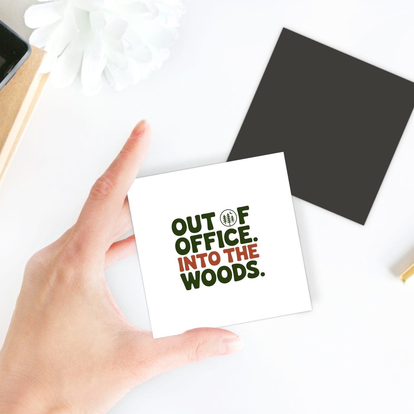 A hand holds the Mirabilia Boutique "Out of Office Into the Woods" magnet—an ideal accent for cabin decor. Nearby are a black card, white flower, and gold-edged notebook, creating a charming scene for any nature lover.