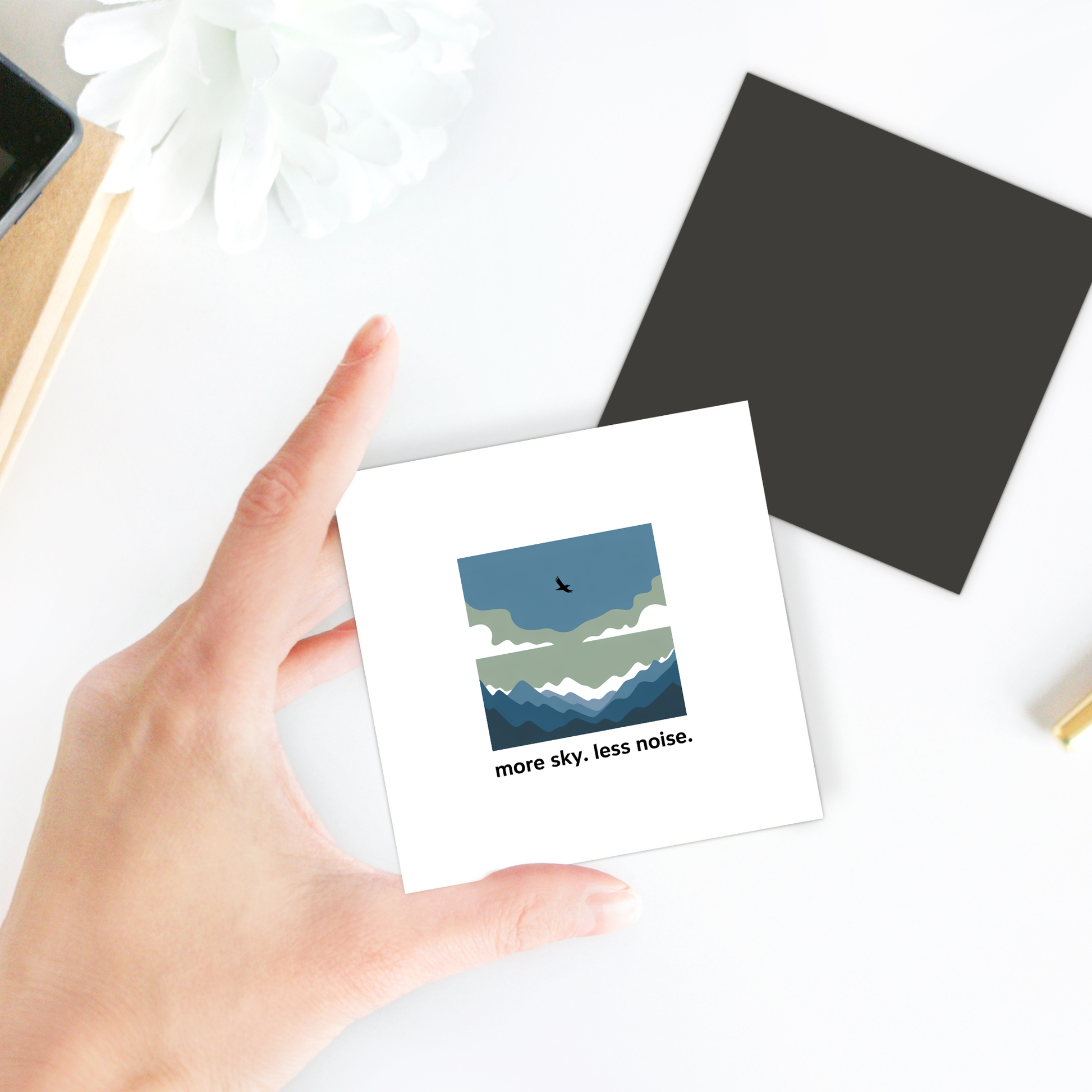 A hand holds the "More Sky Less Noise Nature Magnet" by Mirabilia Boutique, a square decor piece with clouds, mountains, a flying bird, and the quote "more sky. less noise." displayed among other items on a white surface.