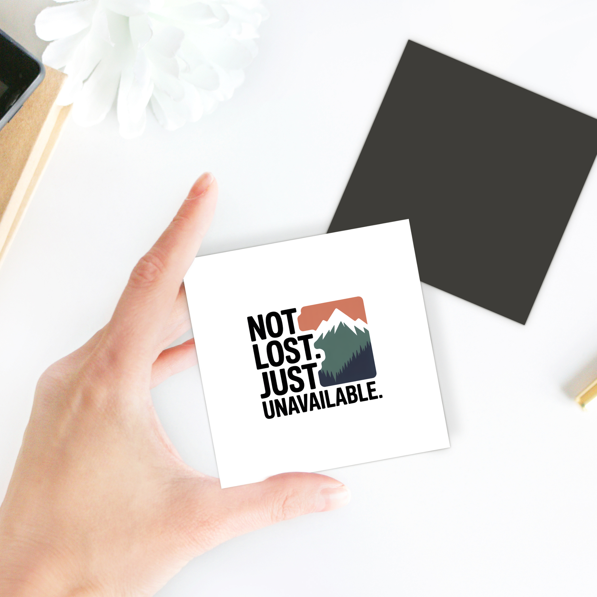 A hand holds the Mirabilia Boutique "Not Lost Just Unavailable Adventure Magnet," featuring a mountain graphic and text, on a white surface with a white flower, black square, and books nearby.