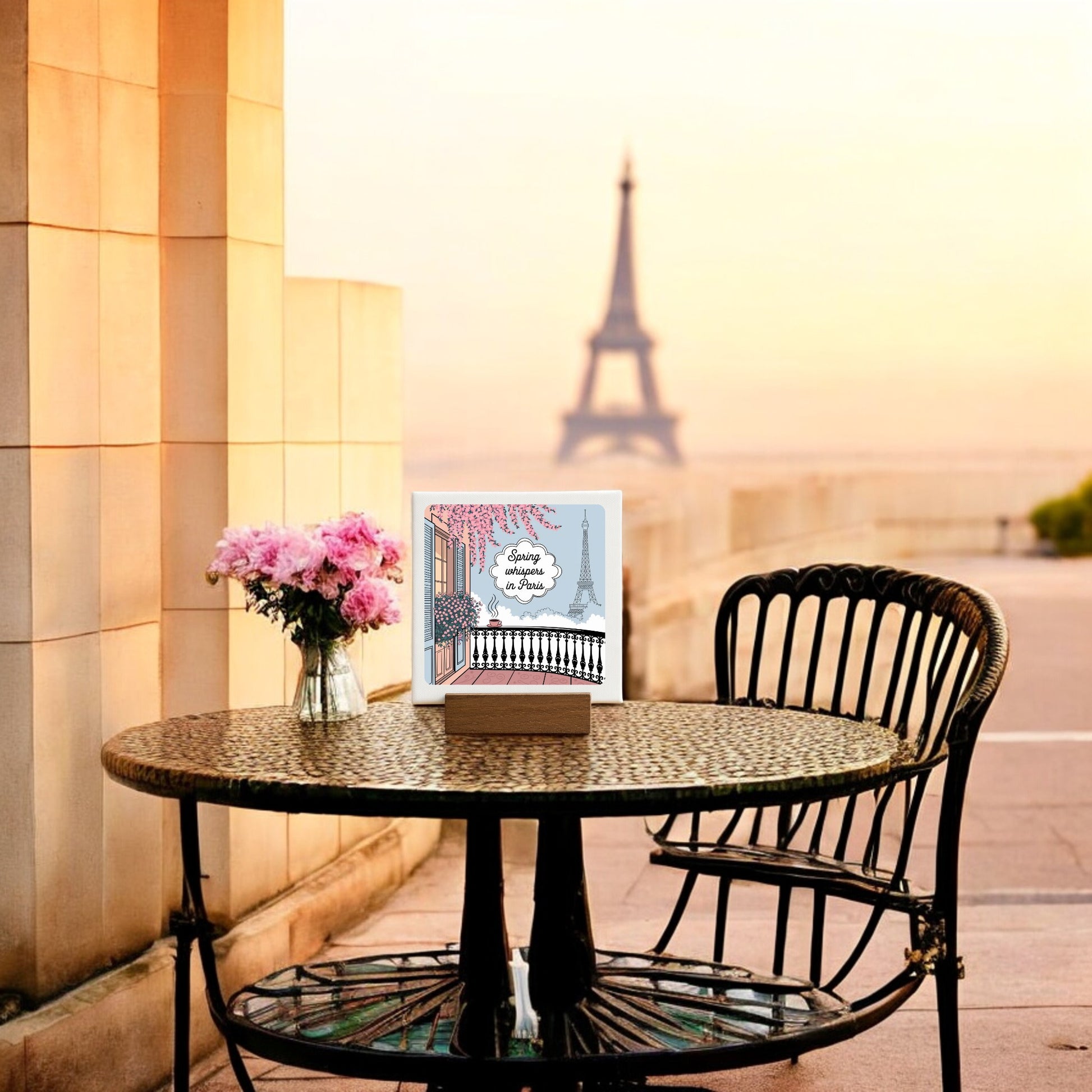 A black chair sits beside a round table with a vase of pink flowers, a book, and the Mirabilia Boutique “Spring Whispers in Paris Mini Sign – Soft Paris Spring,” on a terrace overlooking the Eiffel Tower at sunset in warm, romantic light.