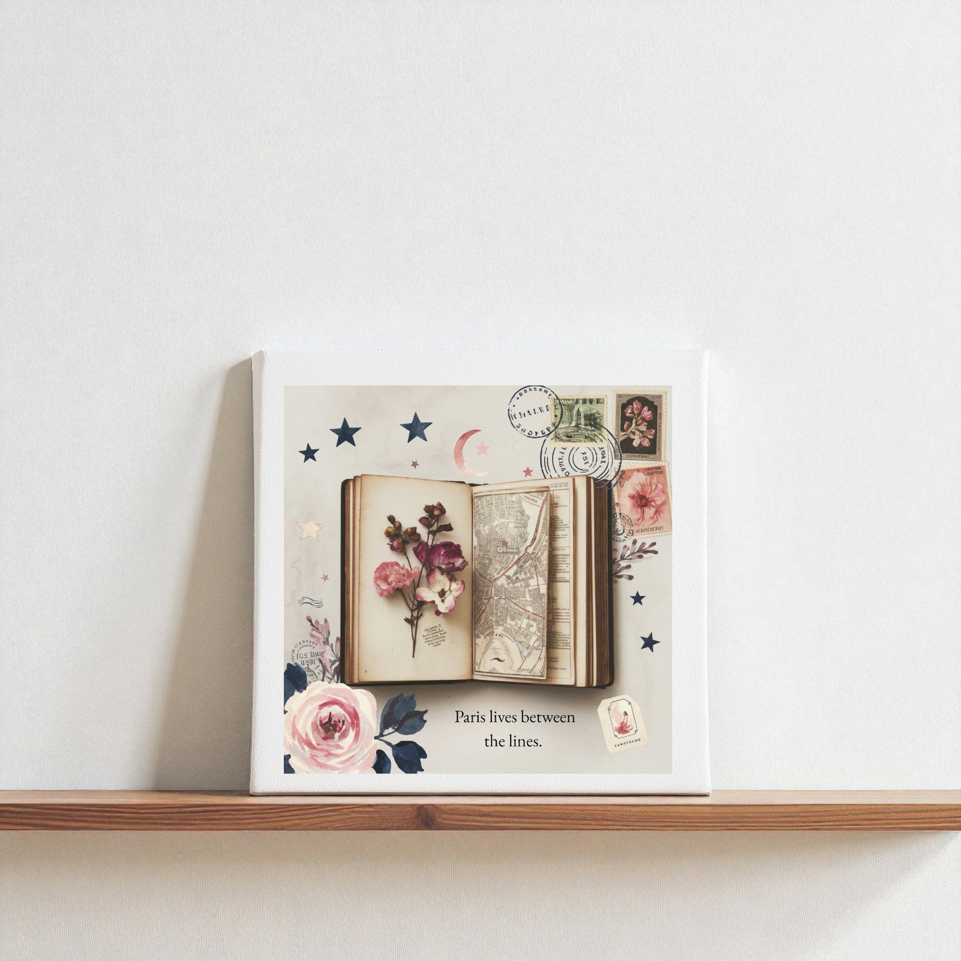 The Paris Between the Lines Canvas – Literary Floral 8x8 Art by Mirabilia Boutique features flowers, a book, a map, stamps, and stars with the caption "Paris lives between the lines"—ideal wall art for literary or Paris lovers.