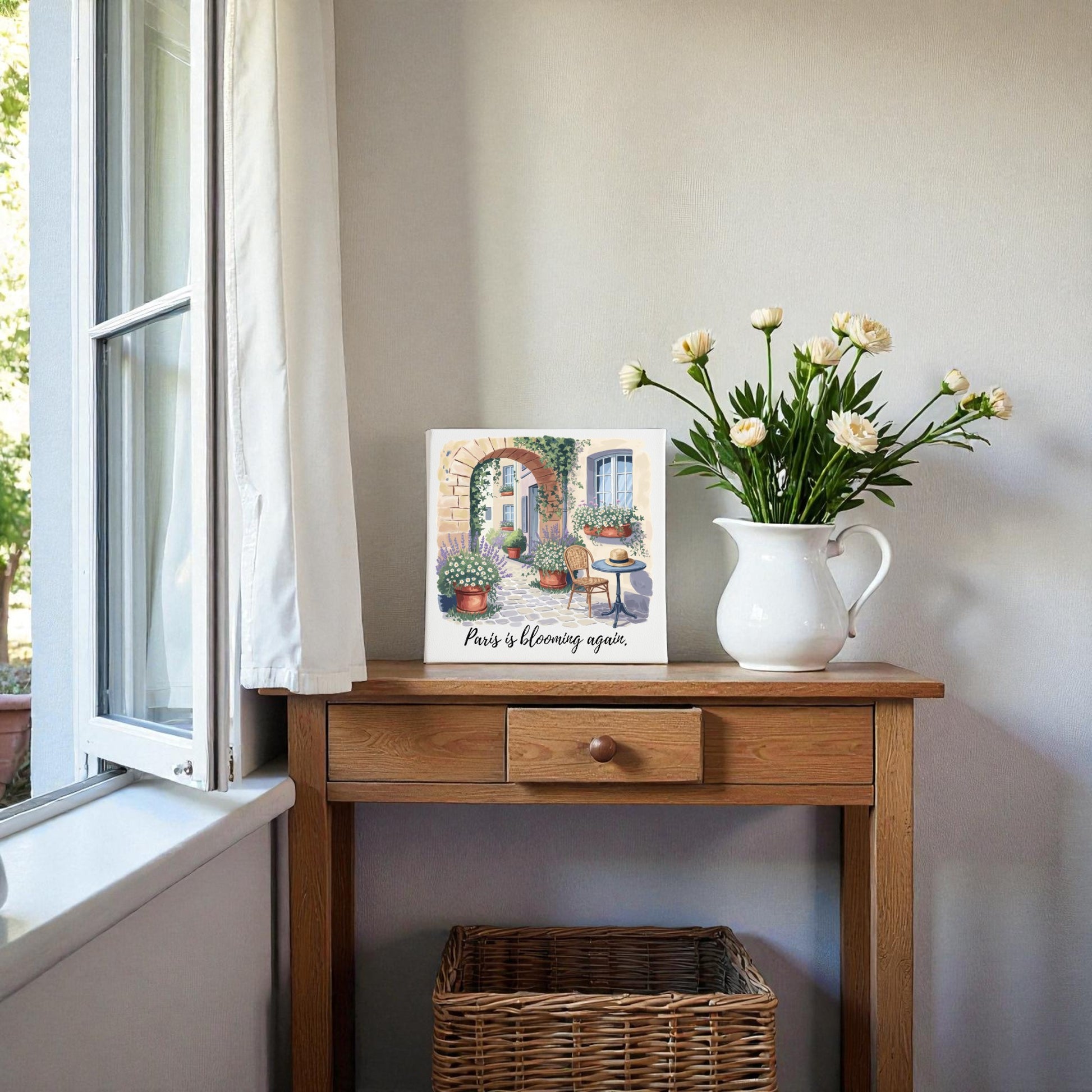 A small wooden table by the window displays a white vase with flowers and the Paris Is Blooming Again Canvas Wall Art by Mirabilia Boutique, bringing French springtime charm. A wicker basket sits below as sunlight streams through the open window.