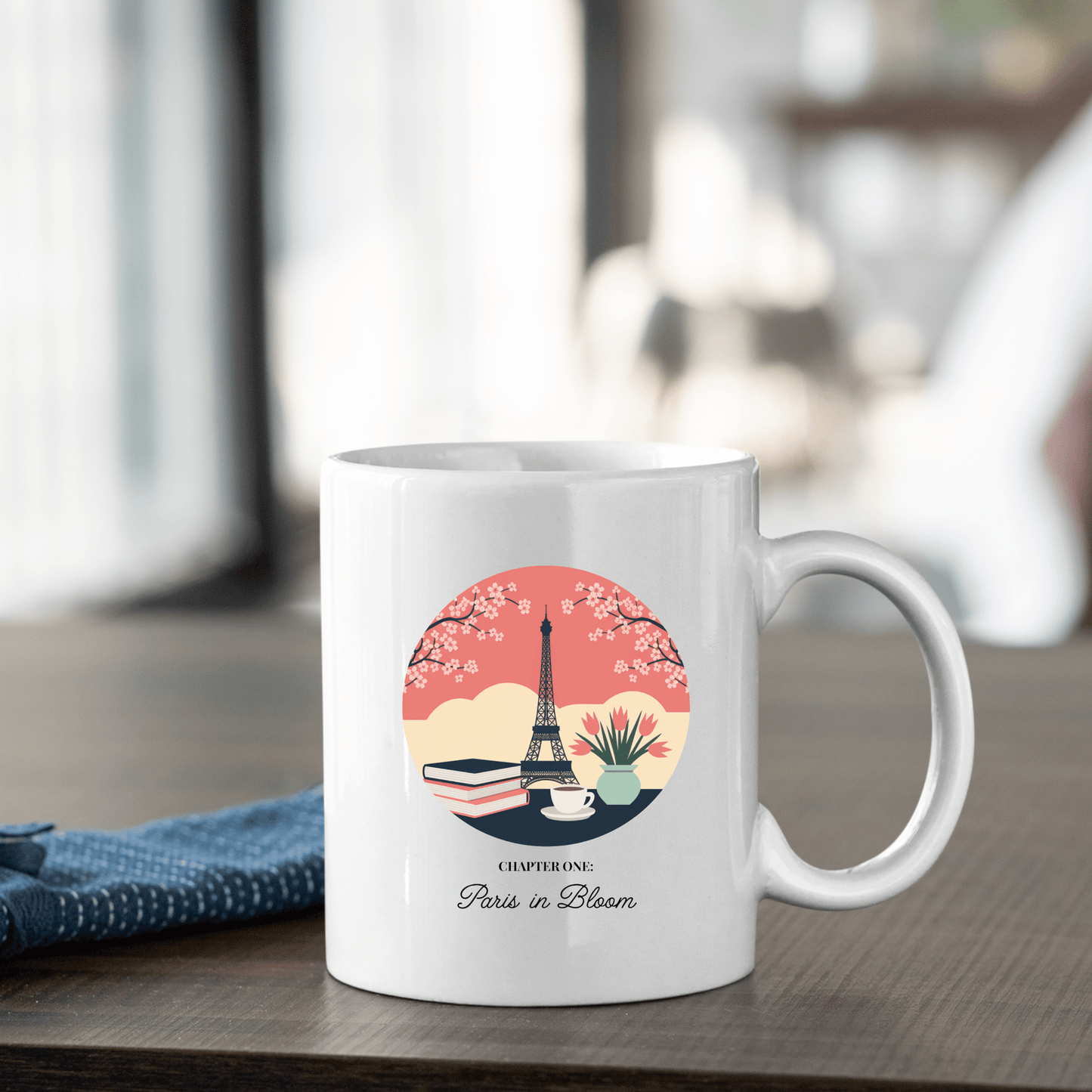 The Paris Book Lover Mug – Chapter One: Paris in Bloom by Mirabilia Boutique sits on a wooden table, featuring the Eiffel Tower, books, flowers, and "CHAPTER ONE Paris in Bloom" text—an ideal gift for book lovers. A blue napkin is nearby.