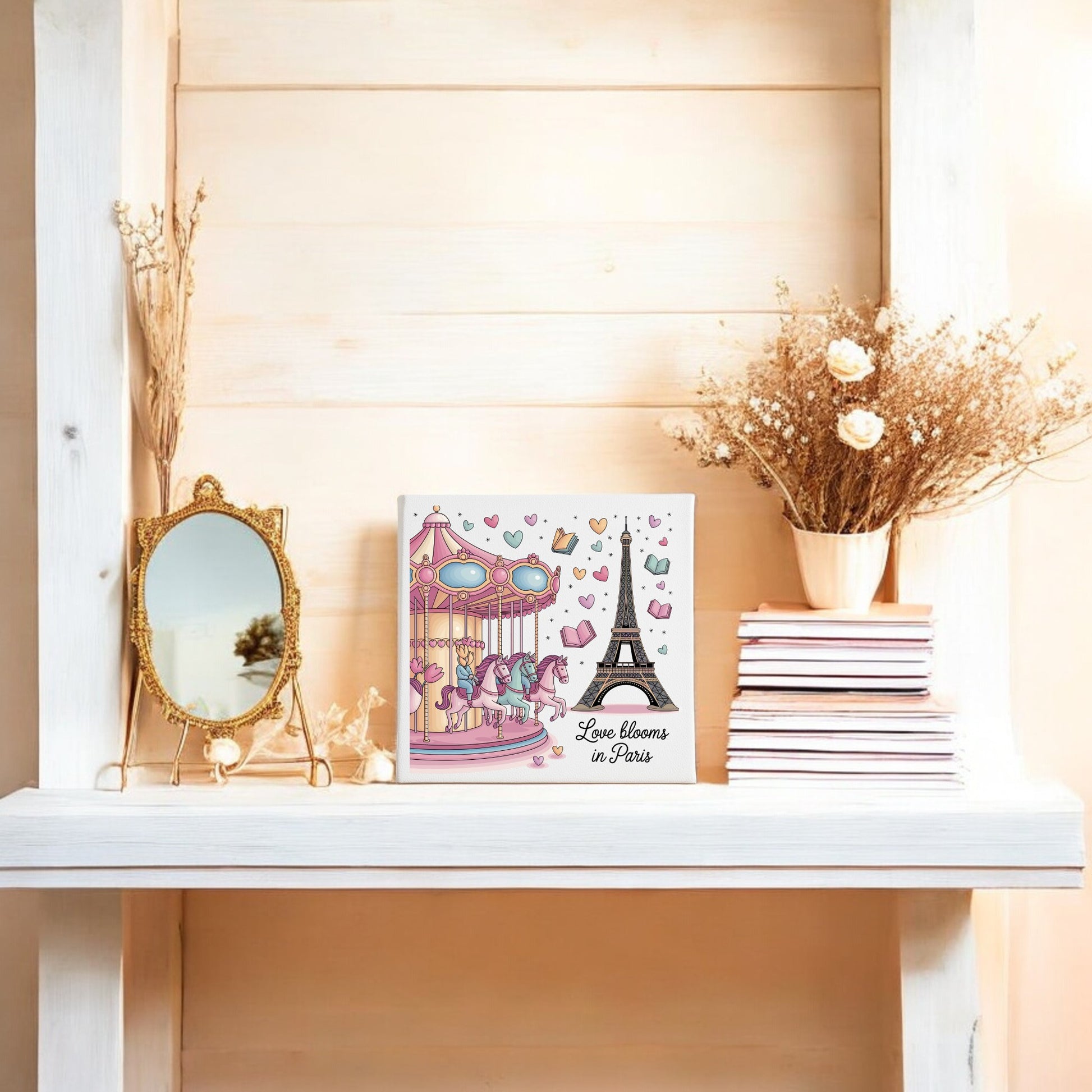 A white shelf displays the "Love Blooms in Paris Canvas – Eiffel Tower & Carousel Art" by Mirabilia Boutique, along with a vintage mirror, pink books, and dried flowers for charming, romantic pastel artwork vibes.