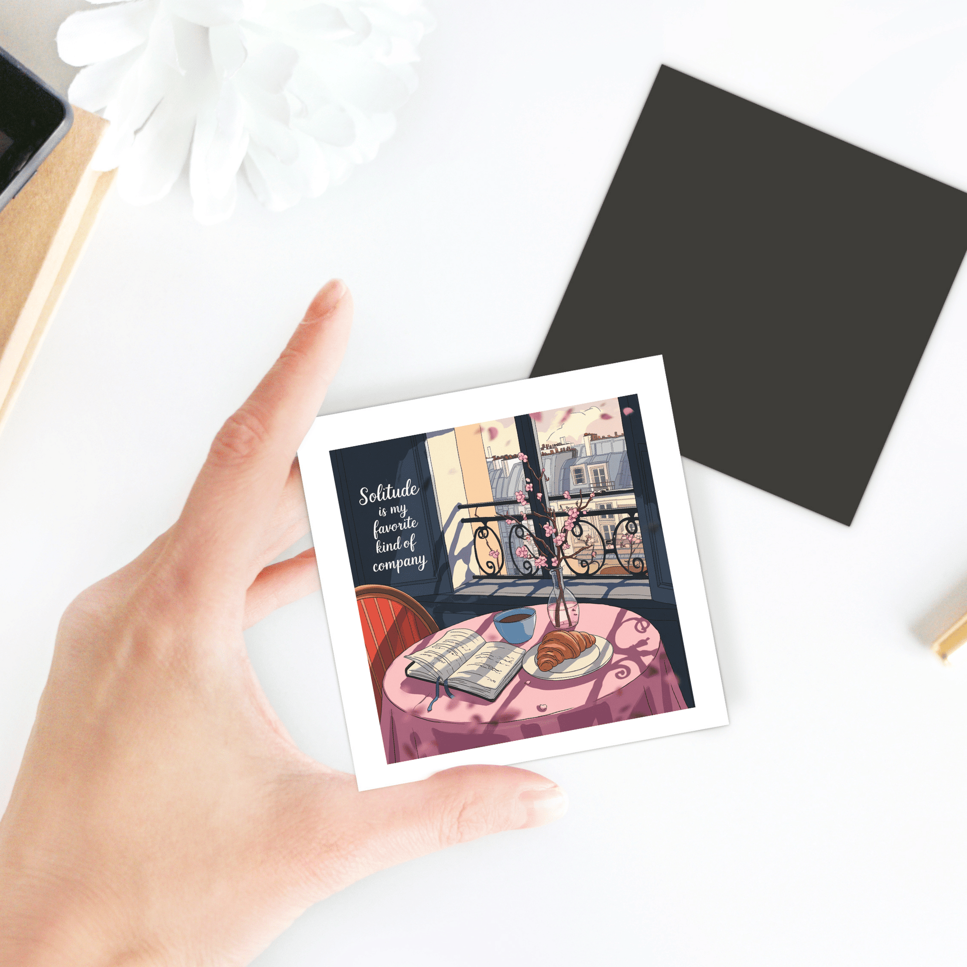 A hand holds the Solitude Magnet – Paris Morning Still Life by Mirabilia Boutique, featuring a cozy table scene with book, coffee, croissant, and city view—Parisian apartment decor. The magnet reads: "Solitude is my favorite kind of company.
