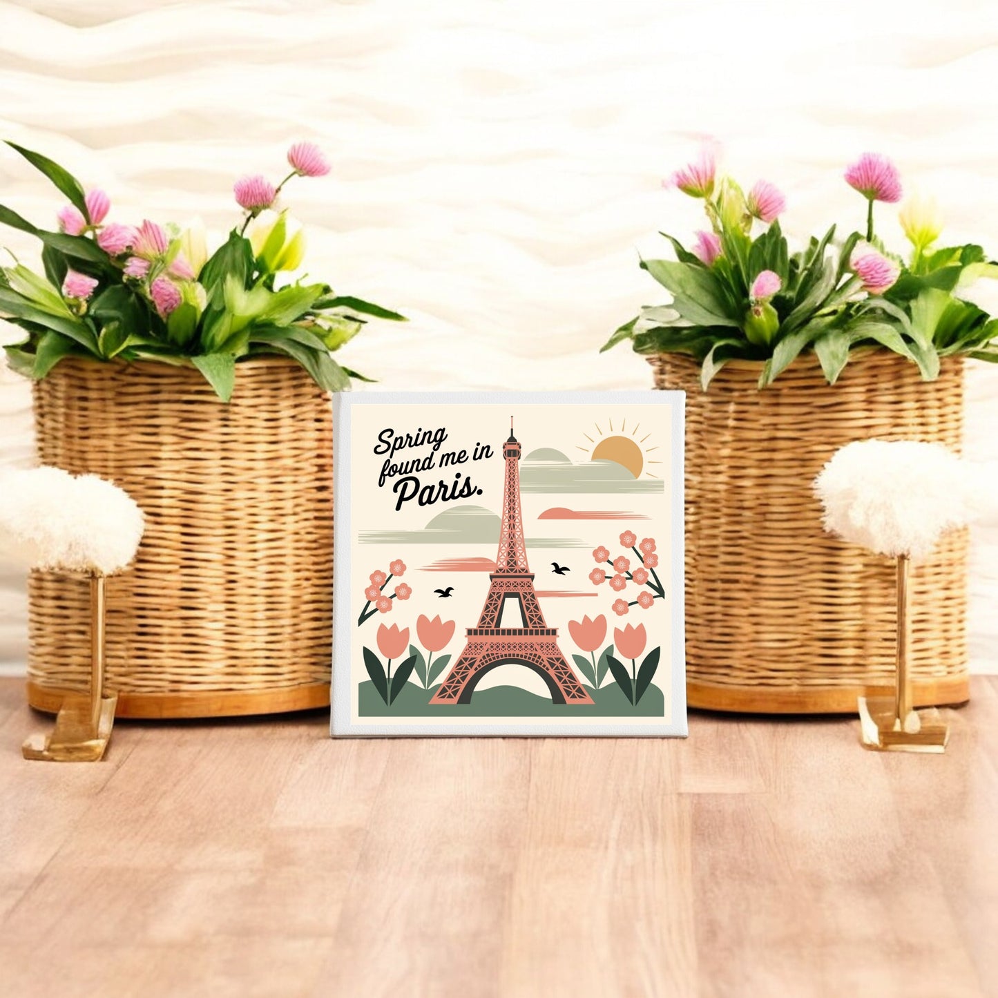 The Mirabilia Boutique "Spring Found Me in Paris – Eiffel Tower Art Canvas" features floral accents and a Parisian motif, displayed on a wooden floor between wicker planters with pink and green blooms against a wavy white wall.