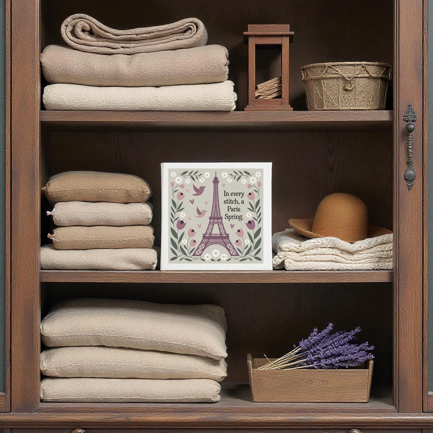 A wooden shelf holds beige towels, a brown hat, and lavender in a tray, complemented by Mirabilia Boutique’s Paris Spring Stitch Canvas—an embroidered-style floral artwork for a soft, natural cottagecore Paris charm.