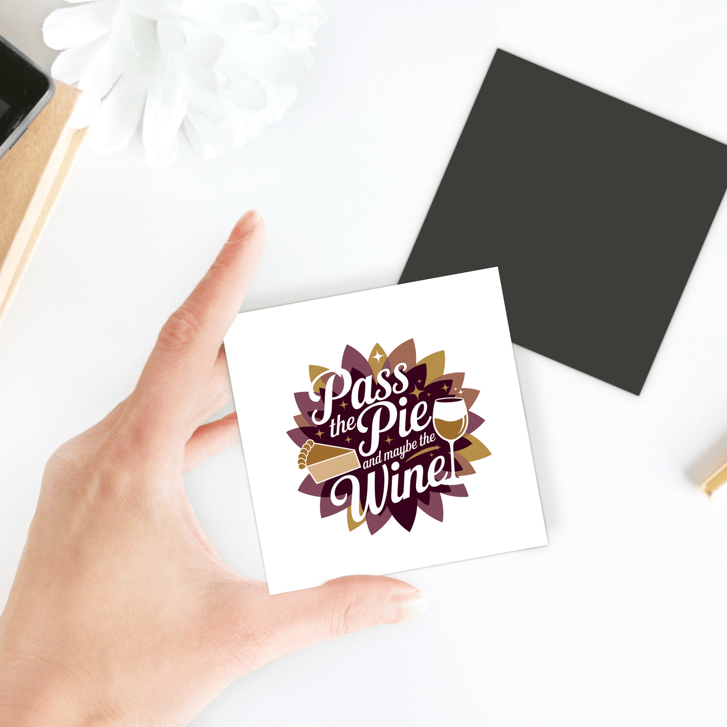 A hand holds the "Pass the Pie & Wine Magnet" by Mirabilia Boutique, a funny Thanksgiving fridge decor gift for wine lovers. It's surrounded by a white flower, black square, and gold pen on a white surface.