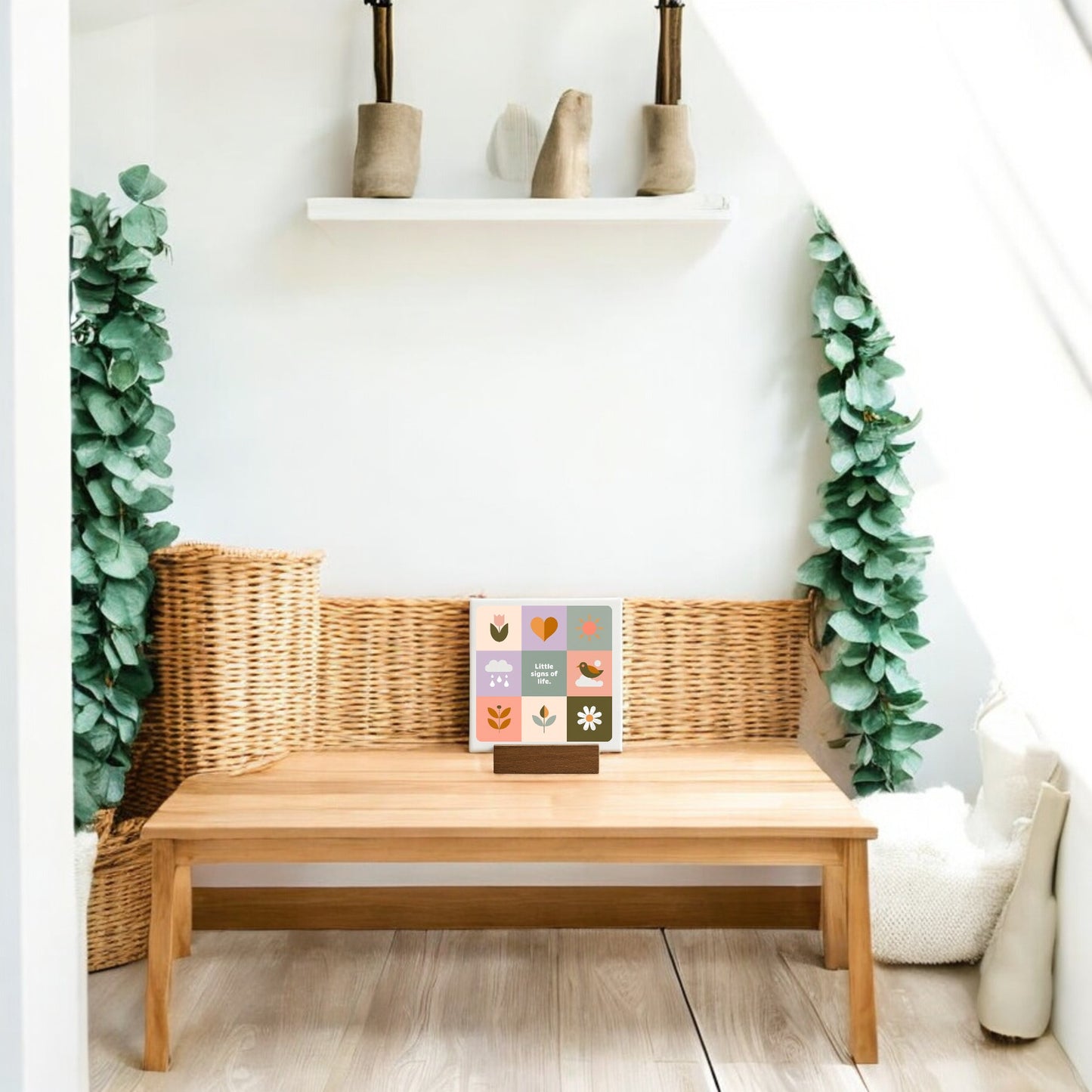 A cozy, minimalist reading nook with a wooden bench, wicker backrest, small table, and greenery. The Little Signs of Life Mini Canvas Sign by Mirabilia Boutique sits beside an illustrated book, adding charm to the softly sunlit space.