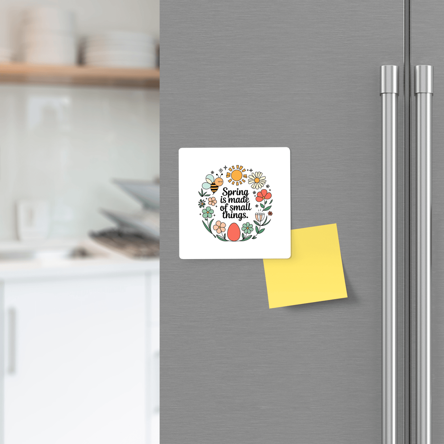 A white card with "Spring is made of small things" is held by the Mirabilia Boutique Spring Is Made of Small Things Floral Magnet on a refrigerator door, next to a yellow sticky note. Blurred kitchen shelves appear in the background.