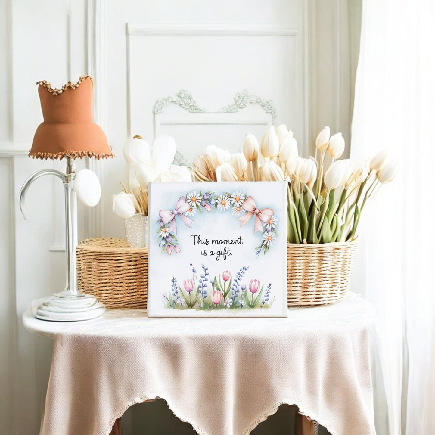 The Mirabilia Boutique "This Moment Is a Gift Canvas" (8x8 spring floral art) is displayed on a beige-draped table with white tulips in wicker baskets, a vintage lamp, and a white-paneled wall for a cozy slow living decor vibe.