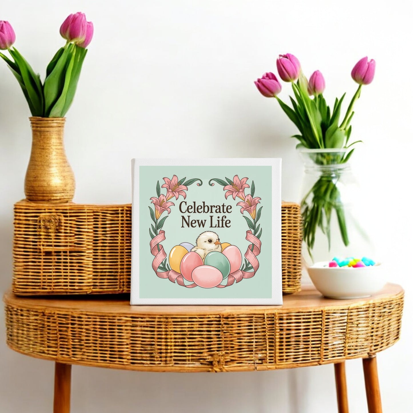 A wicker table displays the "Celebrate New Life Spring Chick Canvas Art" by Mirabilia Boutique—an Easter decoration with a chick, eggs, and flowers—alongside a vase of pink tulips and a bowl of colorful eggs.