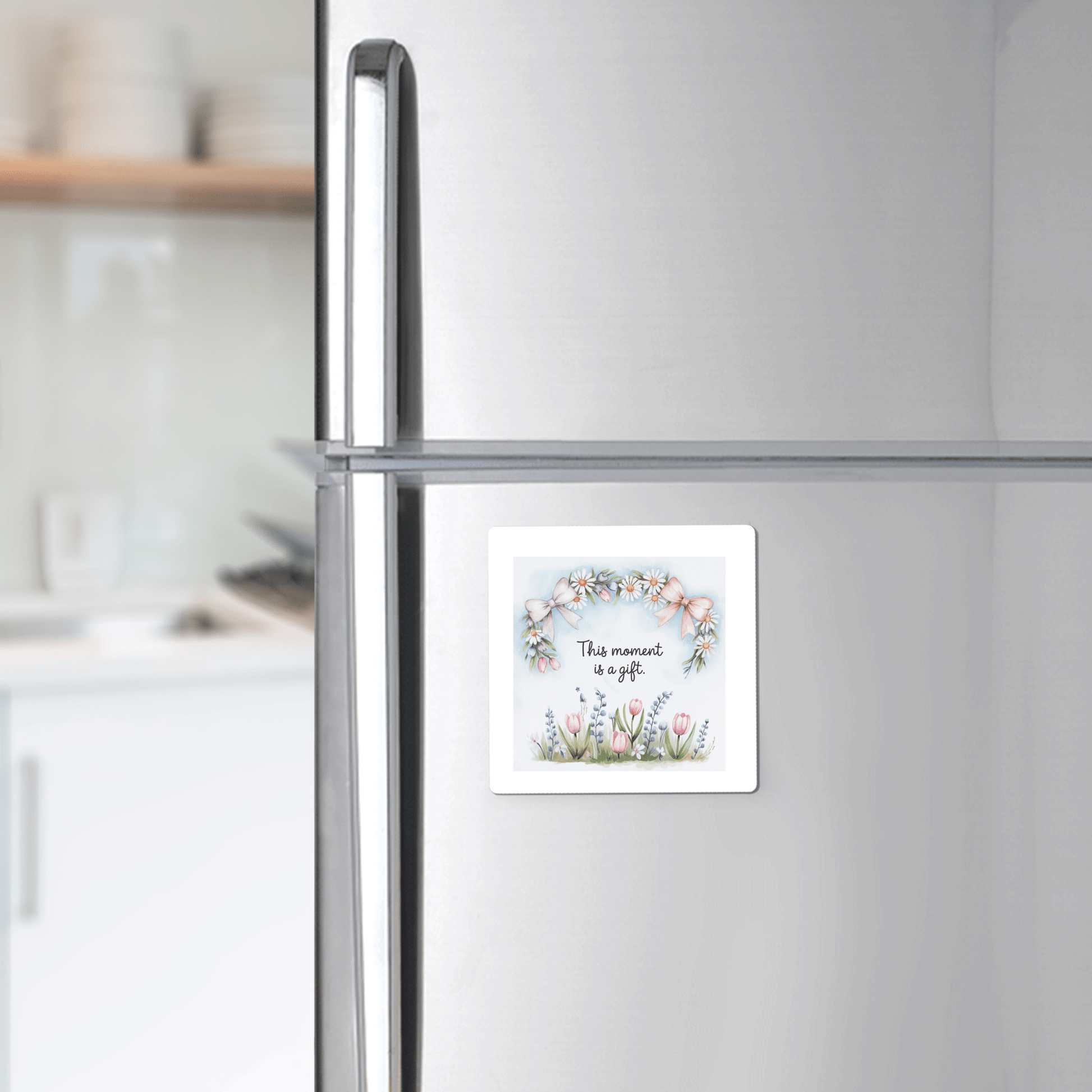 A silver refrigerator displays the Mirabilia Boutique “This Moment Is a Gift Magnet – Floral Spring Quote,” featuring floral art and text. Kitchen shelves with dishes are visible in the background.