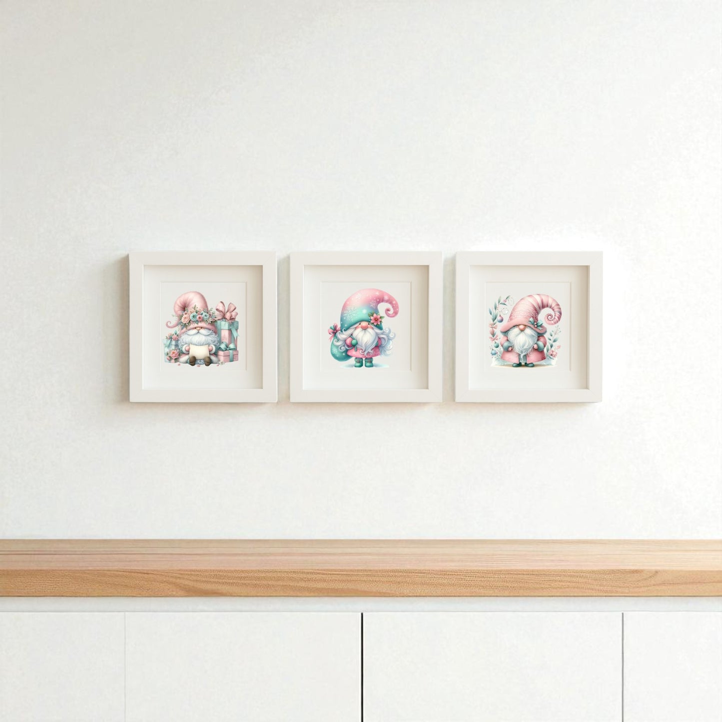 The Pastel Gnome Wall Art Trio by Mirabilia Boutique features three framed prints with pink-hatted gnomes on a clean white wall above a light wood cabinet—perfect for whimsical nursery or winter holiday decor.