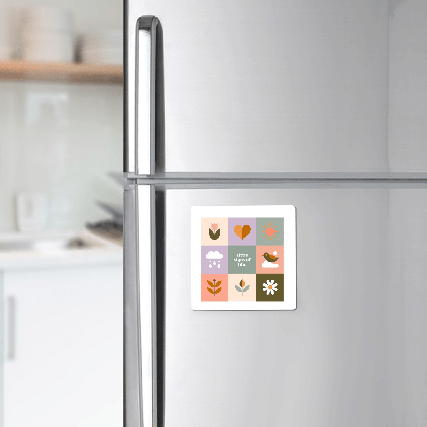 A silver fridge displays the Mirabilia Boutique Little Signs of Life Pastel Grid Magnet, featuring flowers, a heart, bird, sun, and cloud with "Little one." An inspiring gift or minimalist decor accent. A white kitchen is in the background.