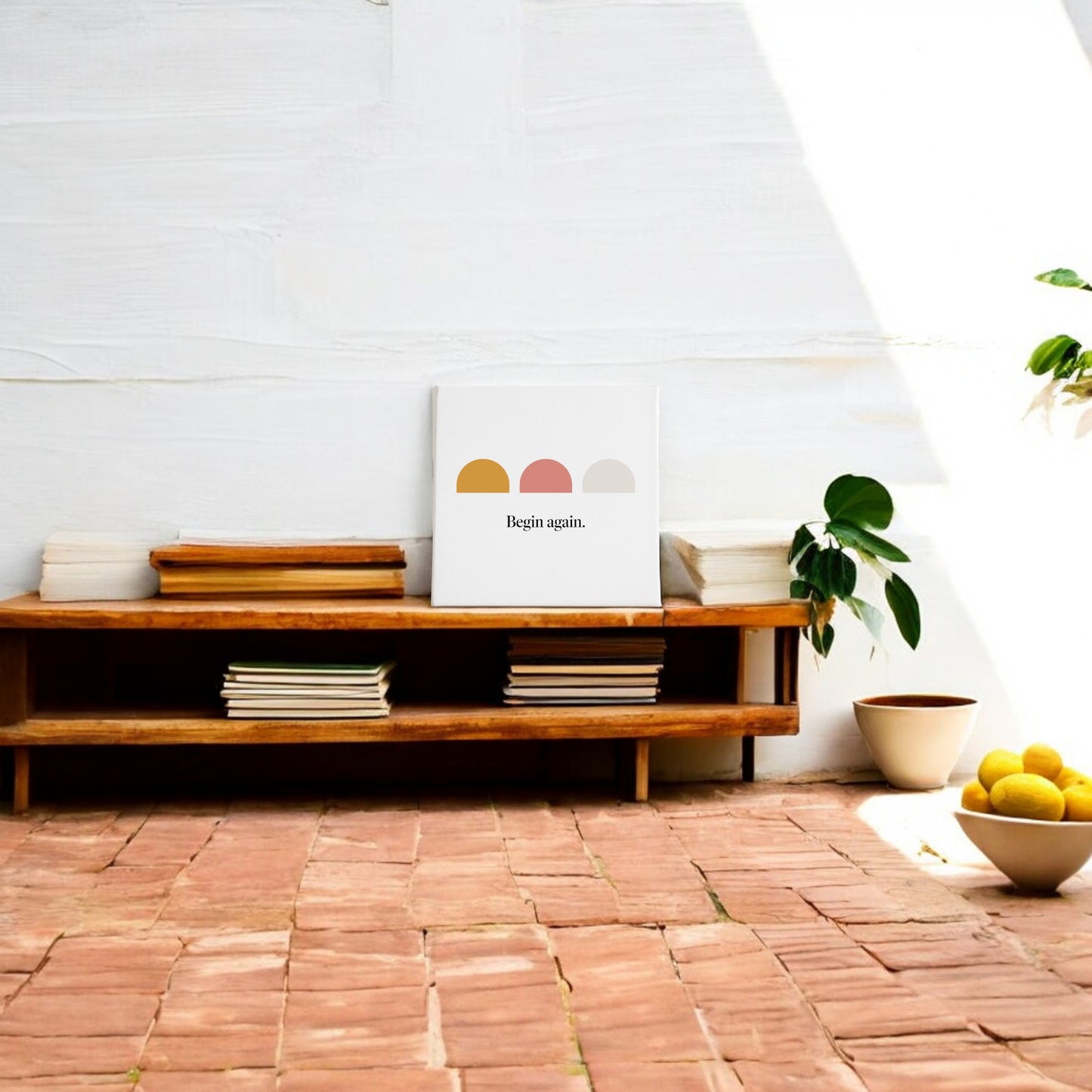 A low wooden bench with stacked books and the Mirabilia Boutique "Begin Again Canvas – Soft Summer Affirmation Wall Art" rests against a white wall. Nearby, a potted plant and a bowl of lemons sit on terracotta tiles in sunlight.