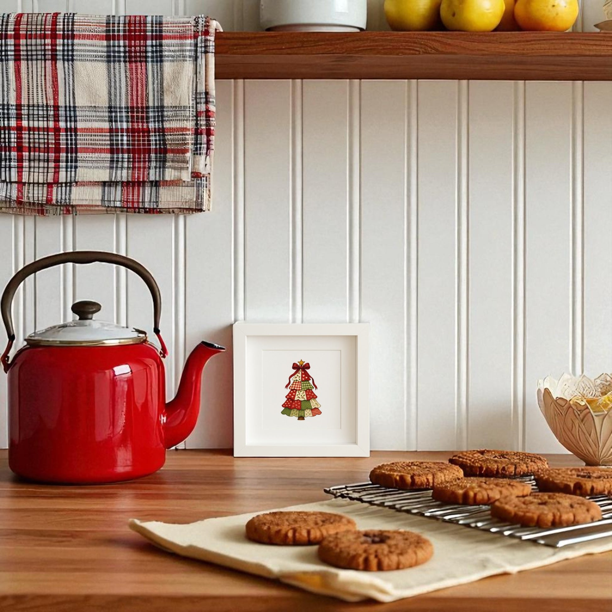 A red kettle sits on a wooden countertop beside the Mirabilia Boutique Patchwork Christmas Tree Sign 5x5 shadowbox. Fresh cookies cool nearby, while a plaid towel and bowl of apples rest on shelves above.
