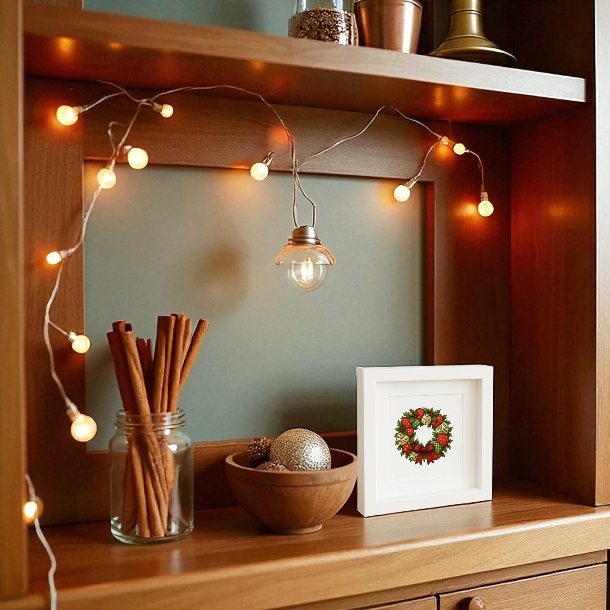 A wooden shelf with string lights, a jar of cinnamon sticks, ornaments in a bowl, and Mirabilia Boutique’s Patchwork Christmas Wreath Sign (5x5 Holiday Shadowbox Art) creates cozy farmhouse holiday decor and a festive atmosphere.