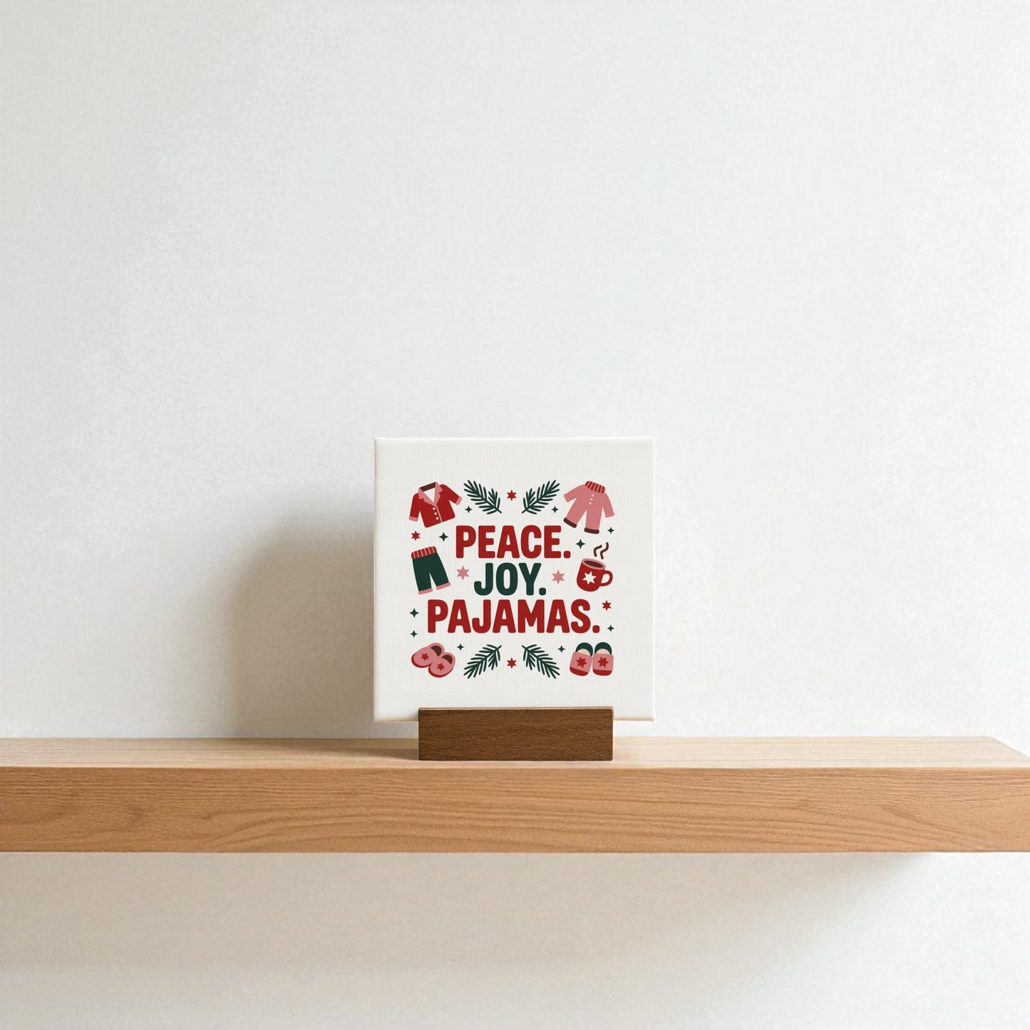 The Mirabilia Boutique Peace Joy Pajamas Mini Sign (4x4) sits on a wooden shelf, featuring red text and festive icons—pajamas, slippers, stars, and evergreen—making it perfect holiday decor against a plain white wall.