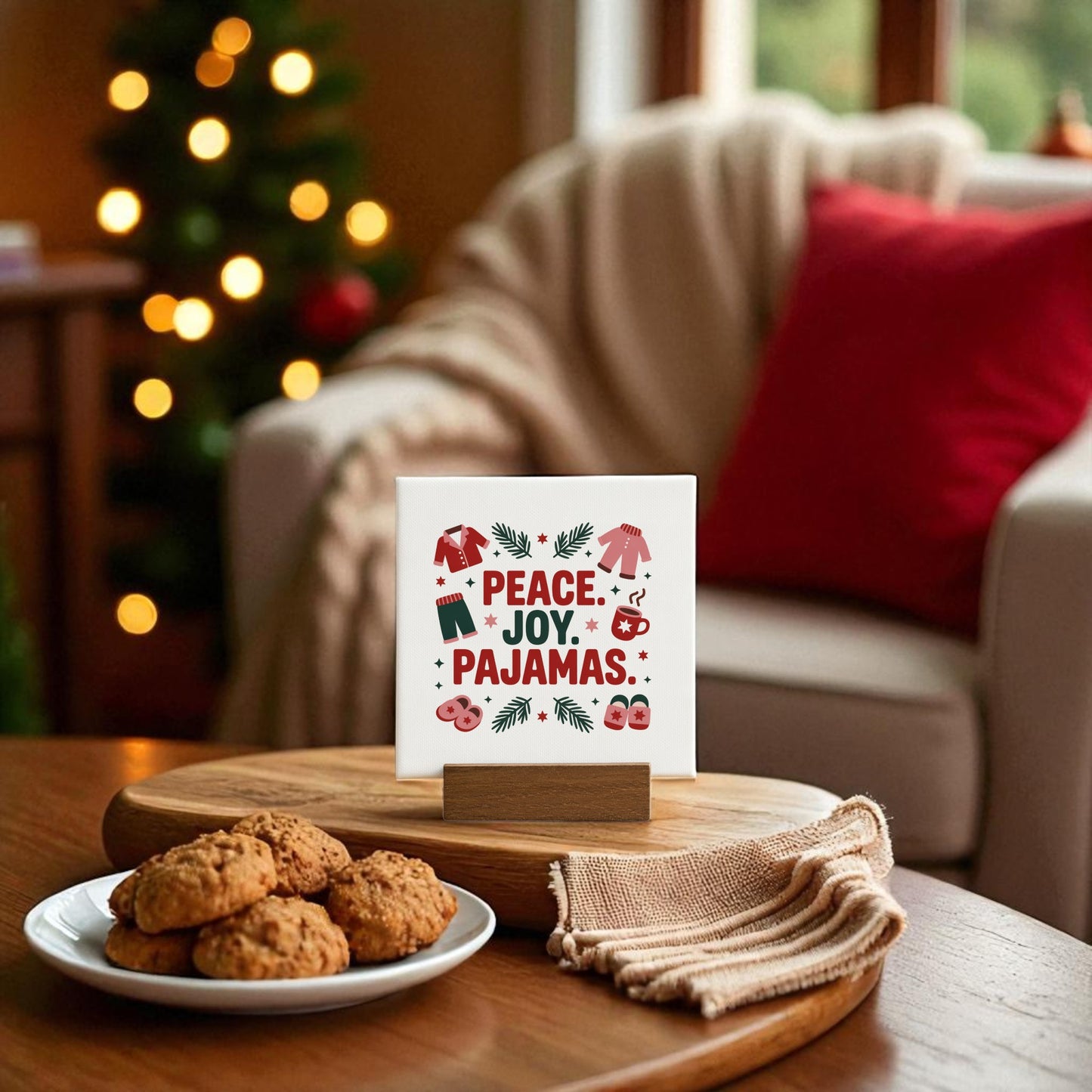 A holiday living room with cookies and a napkin on the table features the Mirabilia Boutique Peace Joy Pajamas Mini Sign (4x4 Holiday Decor Gift) amid festive Christmas decor, a blurred tree, and a cozy chair with a red pillow.