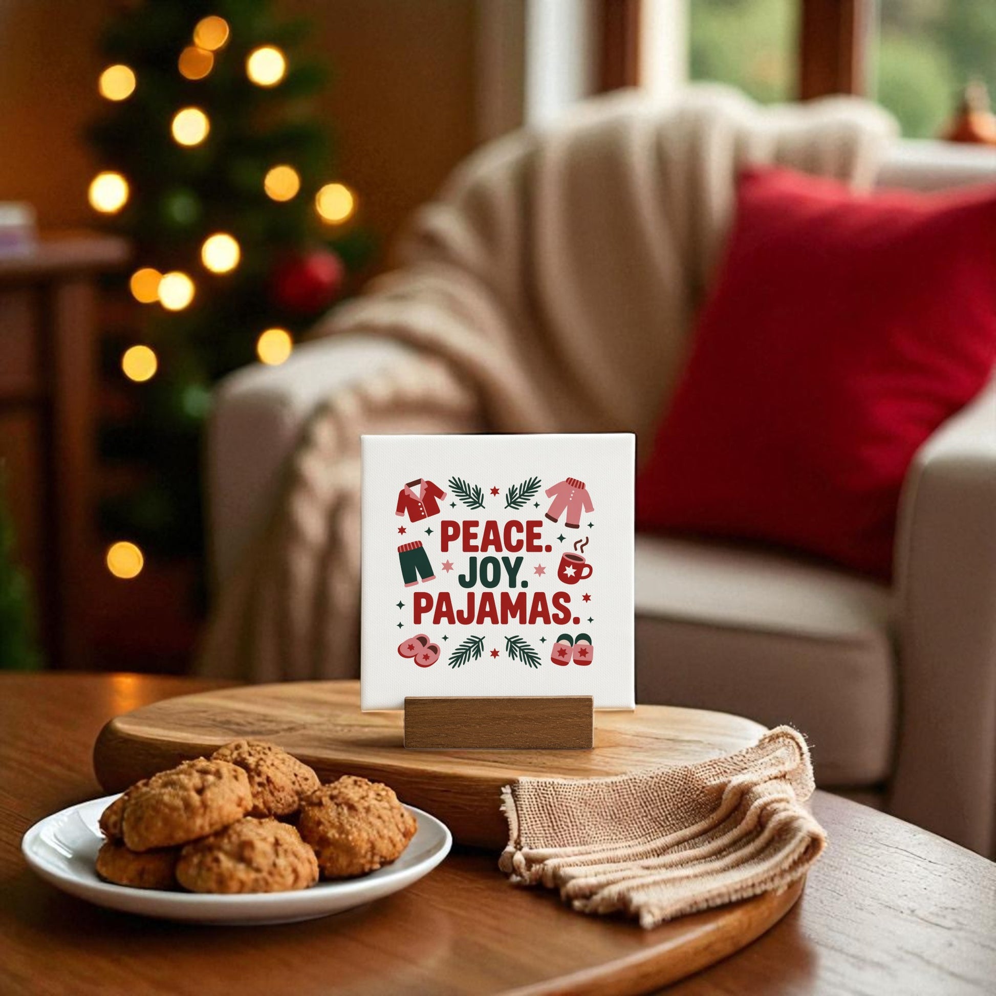 A holiday living room with cookies and a napkin on the table features the Mirabilia Boutique Peace Joy Pajamas Mini Sign (4x4 Holiday Decor Gift) amid festive Christmas decor, a blurred tree, and a cozy chair with a red pillow.