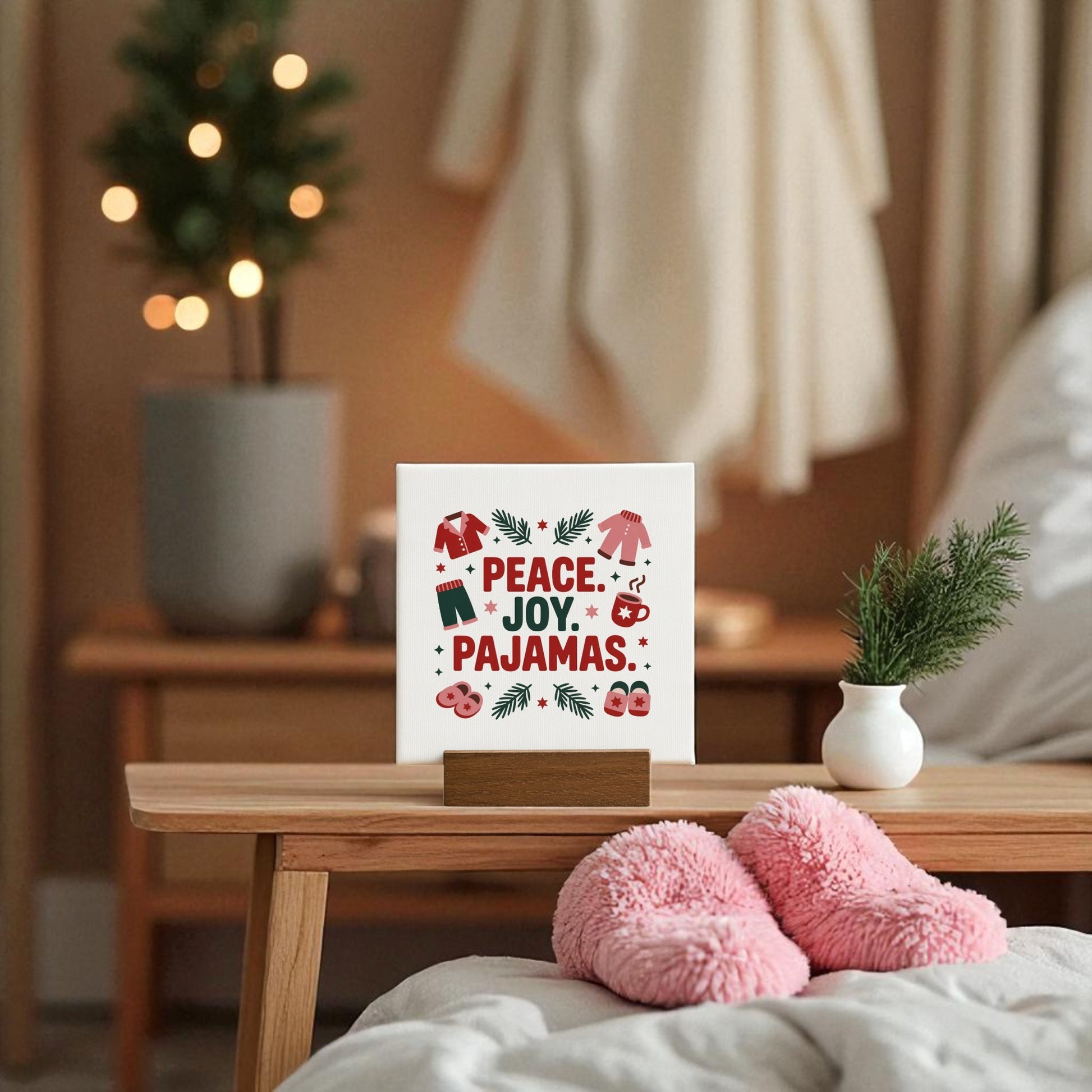 A cozy bedroom displays the Mirabilia Boutique Peace Joy Pajamas Mini Sign (4x4 holiday decor gift) on a wooden table, with pink slippers, a small potted plant, and blurred Christmas tree in the background.