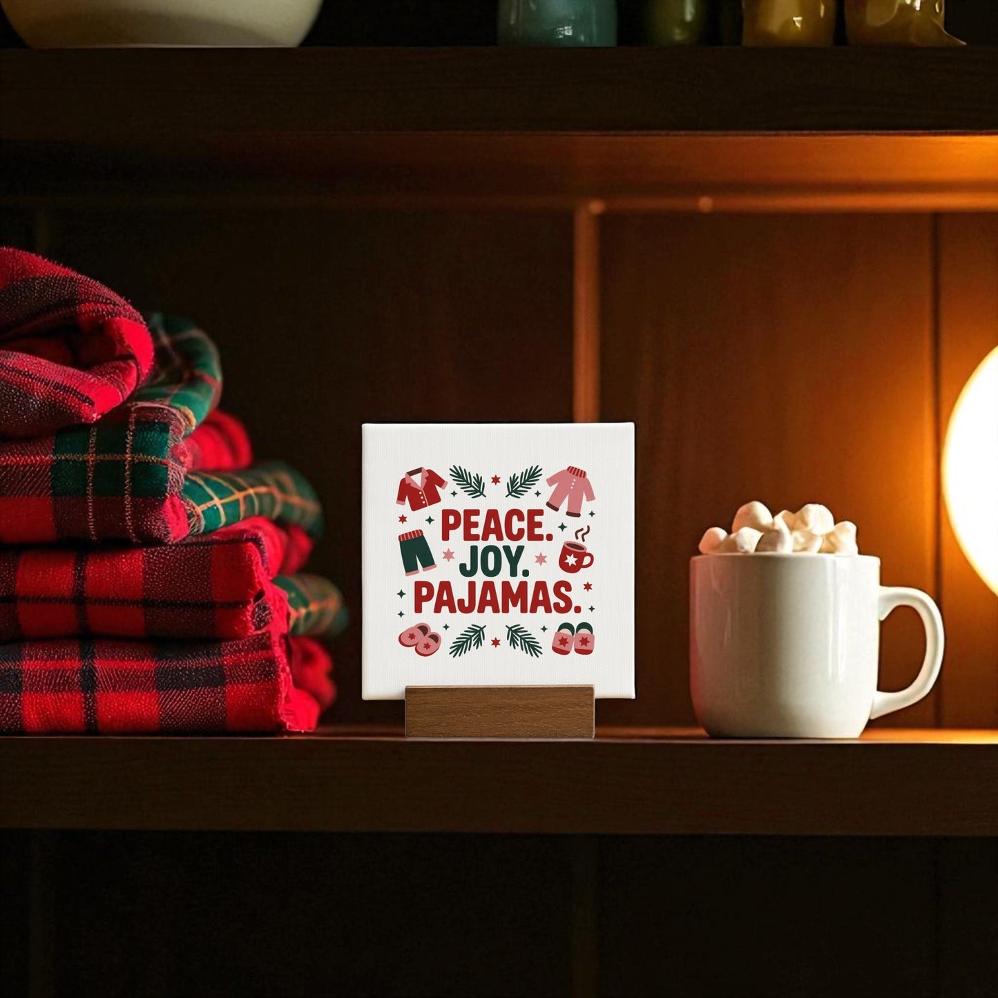 Folded plaid pajamas, a mug of hot chocolate with marshmallows, and Mirabilia Boutique’s “Peace Joy Pajamas” 4x4 holiday mini sign are displayed on a wooden shelf under a warm lamp.