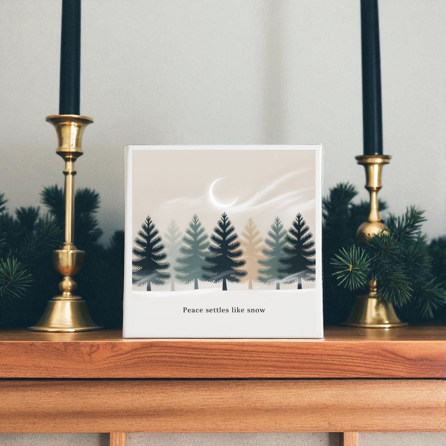 A mantel adorned with greenery and brass candlesticks displays the "Peace Settles Like Snow Canvas" by Mirabilia Boutique—winter minimalist wall art featuring pine trees, a crescent moon, and serene text for nursery decor.