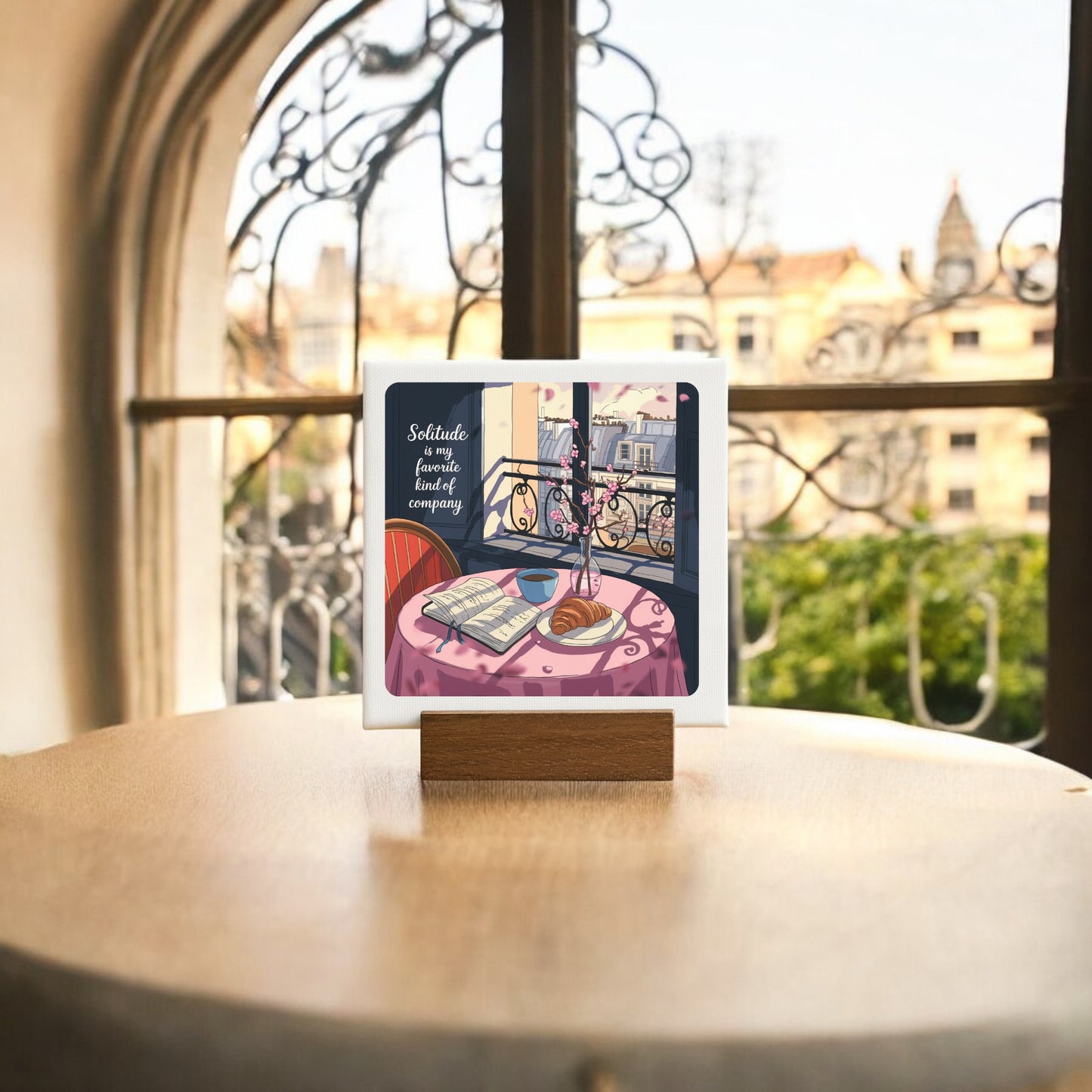 The Solitude Mini Sign – Parisian Morning Still Life by Mirabilia Boutique features a framed table scene with a book, coffee, and croissant on a Parisian balcony—ideal for balcony decor or as a thoughtful gift. Blurred cityscape and arched windows complete the look.