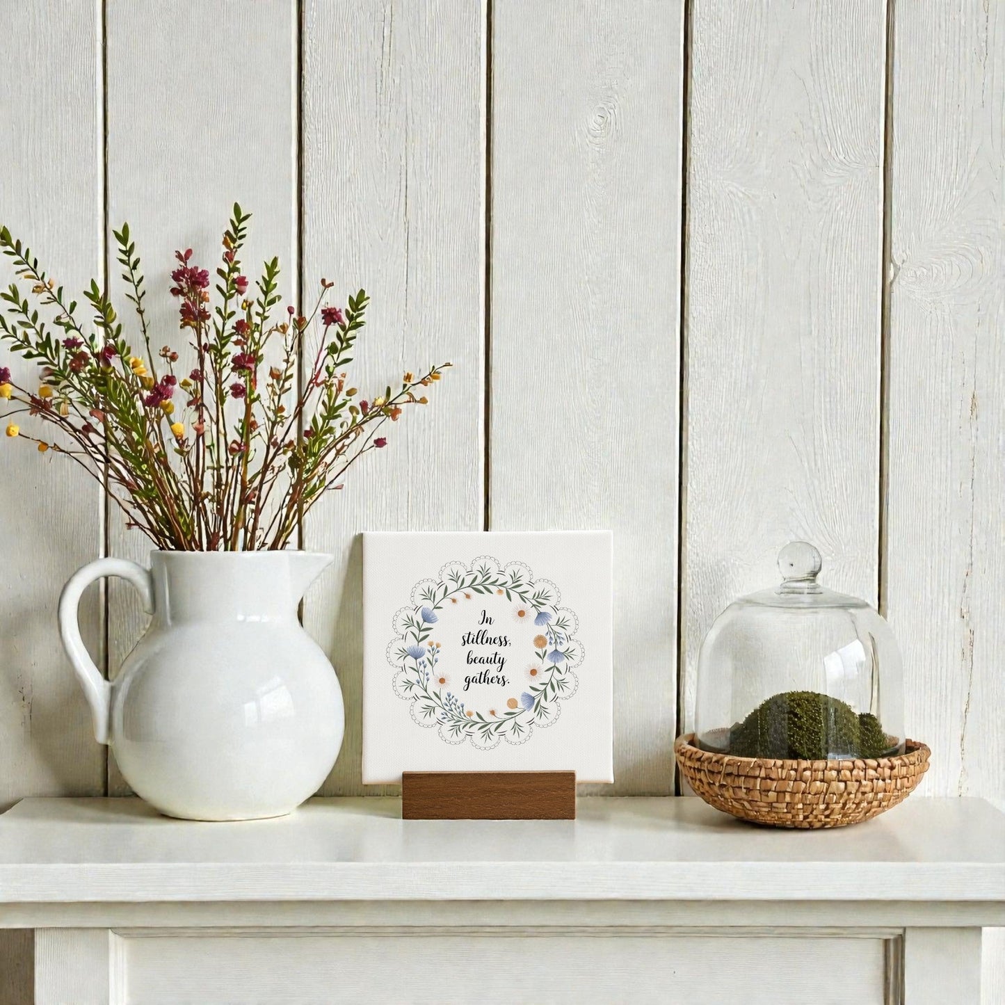 A Mirabilia Boutique "In Stillness Beauty Gathers" mini canvas sign sits on a white meditation shelf with a white pitcher of wildflowers, framed floral art, and a glass cloche over moss atop a woven tray.