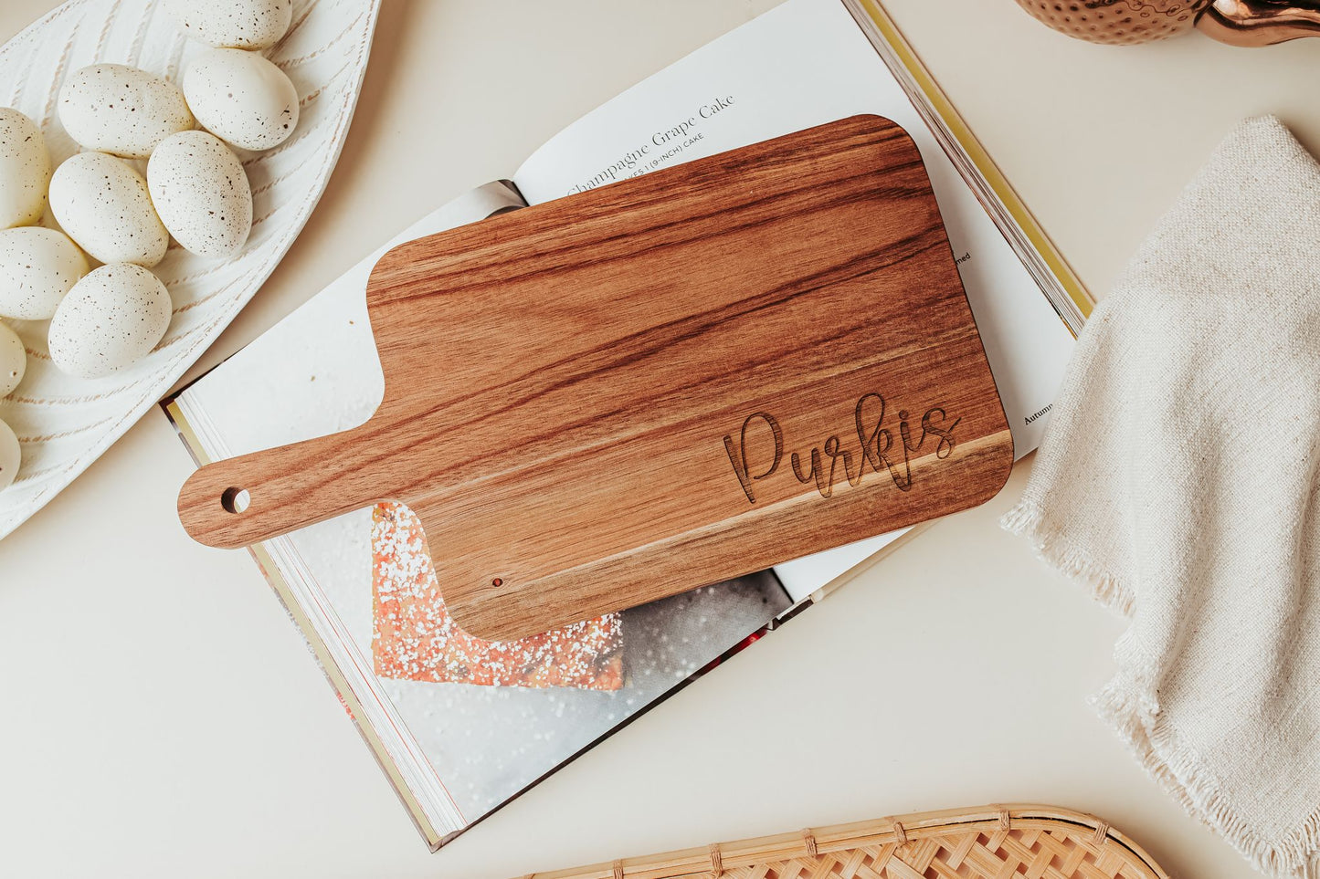 The Engraved Acacia Cutting Board by Mirabilia Boutique (14x8) features "Purples" and rests on an open cookbook, with a towel, eggs, and a woven tray nearby—a perfect custom personalized gift for any occasion.