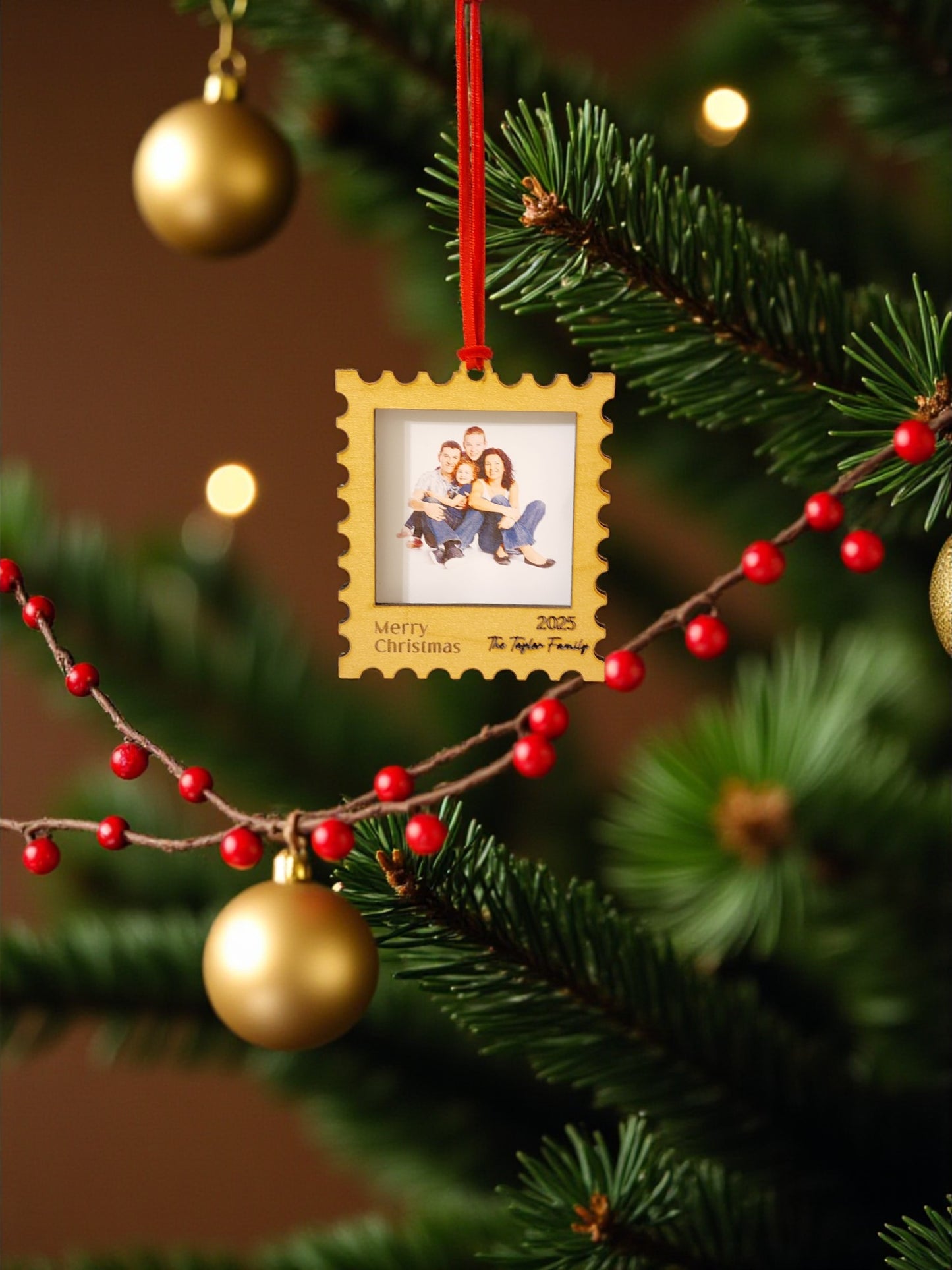 A close-up of a Christmas tree branch adorned with red berries, gold baubles, and the Mirabilia Boutique Personalized Christmas Stamp Frame Ornament featuring a family photo and "Merry Christmas 2023 The Taylor Family" text.