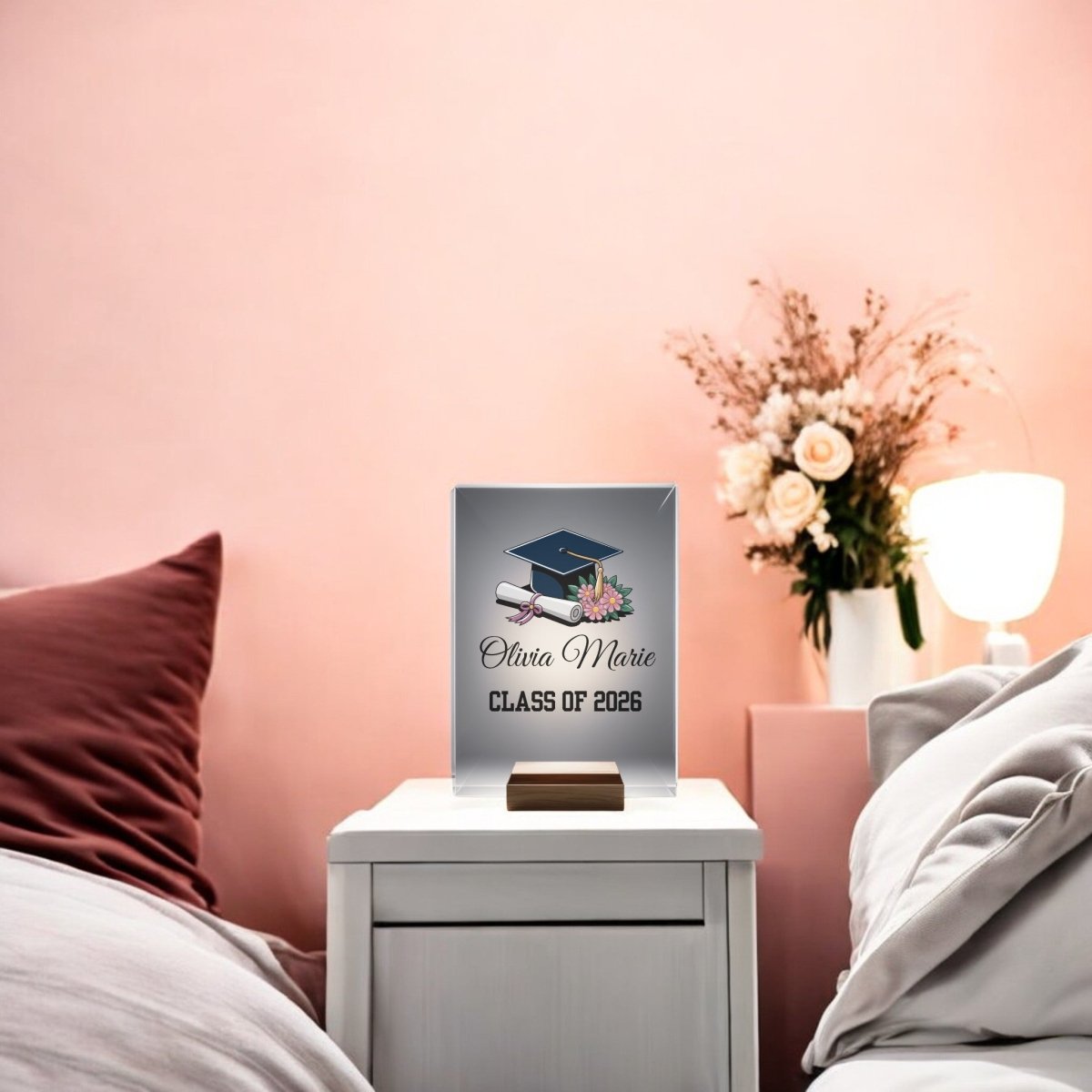 A Floral Personalized Graduation Night Light by Mirabilia Boutique sits on a white bedside table, displaying “Olivia Marie, Class of 2026” with a graduation cap and diploma; a lamp and flowers decorate the pink-walled background.