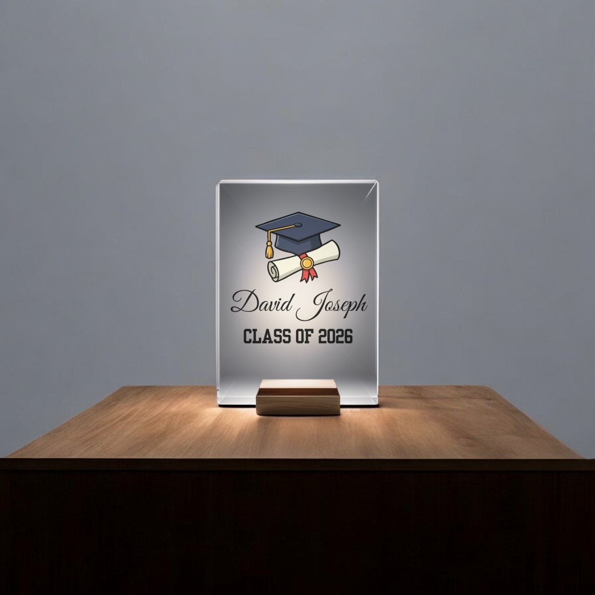 The Mirabilia Boutique Classic Graduation Cap Personalized Night Light features a glass plaque on a wooden base, illuminated from below, with a graduation cap, diploma, and "David Joseph CLASS OF 2026" engraved.