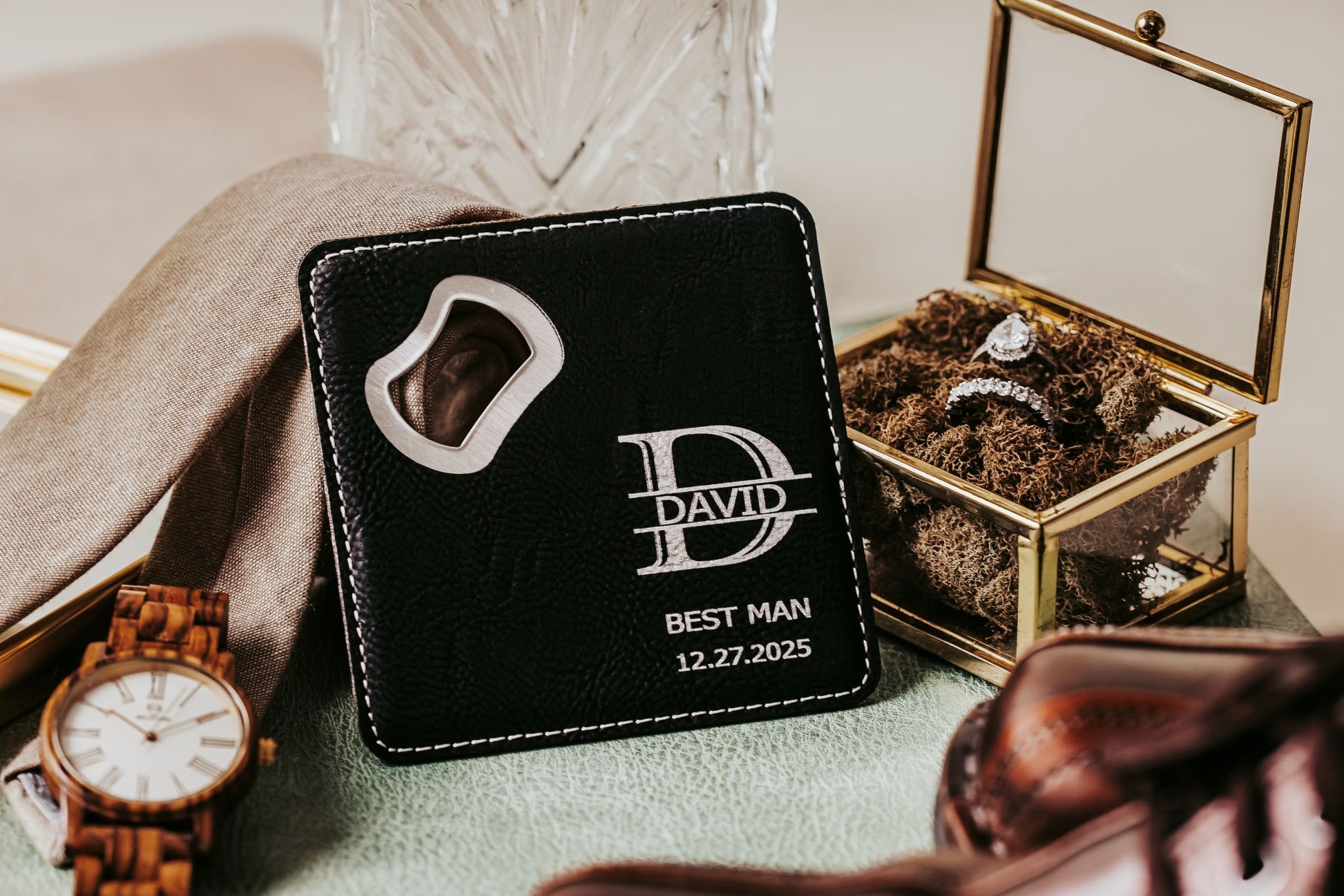 The Mirabilia Boutique Engraved Leatherette Bottle Opener Coaster, featuring "David, Best Man, 12.27.2025," rests beside a watch, tie, ring box, and shoes—a thoughtful personalized best man gift for a special occasion.