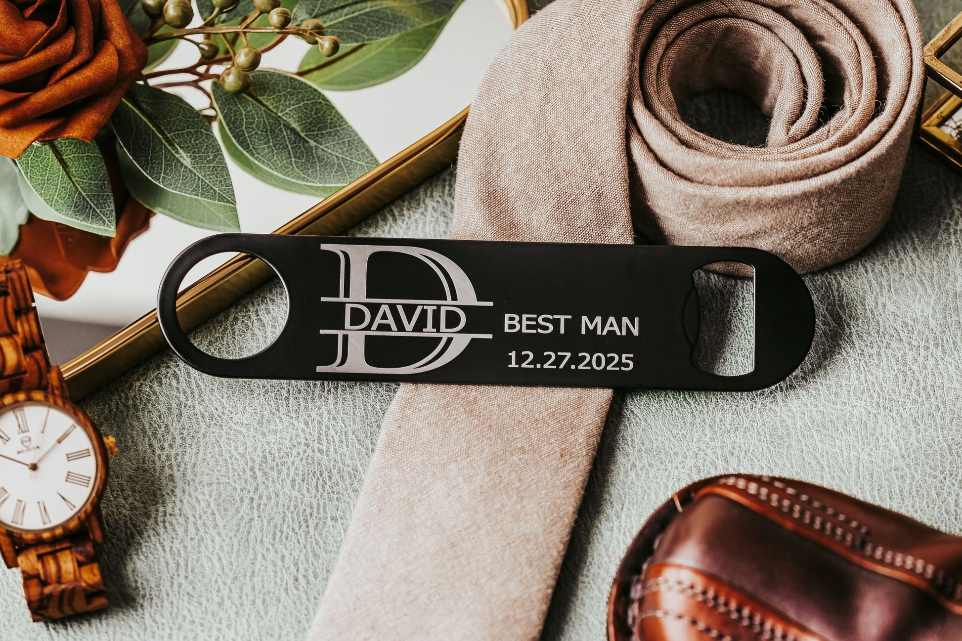 The Engraved Flat Metal Bottle Opener by Mirabilia Boutique, personalized with “DAVID BEST MAN 12.27.2025”, sits on a beige tie among classic gifts—an ideal best man present and memorable groomsmen favor.
