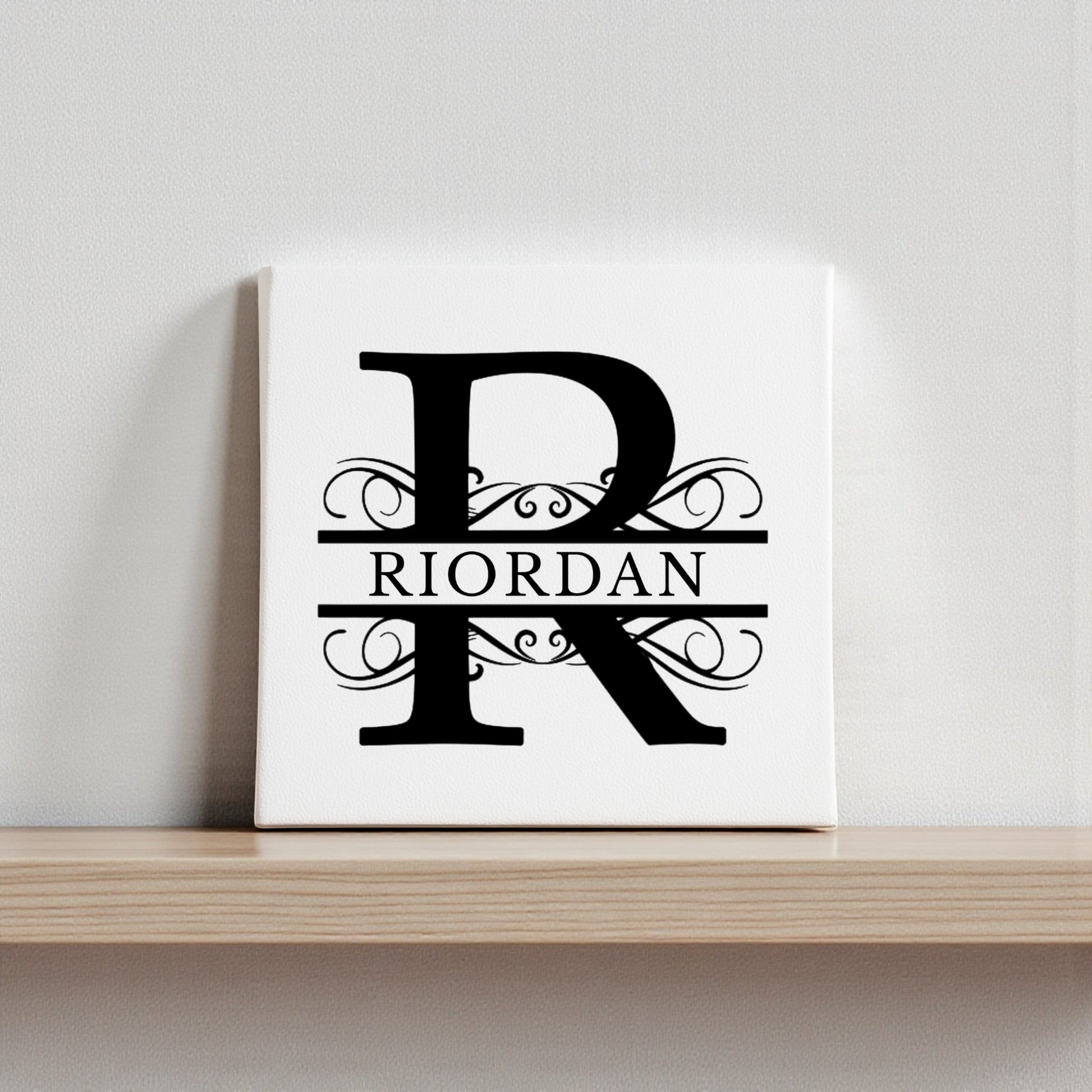 The Mirabilia Boutique Custom Monogram Canvas with Family Name – 8x8 showcases a bold black "R" with ornate swirls and "RIORDAN" across the center, creating elegant, personalized wall art for your home.