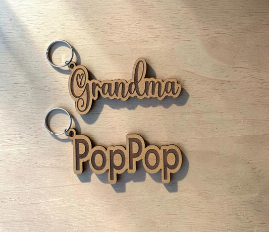 Two Personalized Engraved Wood Name Keychains from Mirabilia Boutique rest on a light wood surface. One keychain features "Grandma" in elegant cursive script, while the other displays "PopPop" in bold, rounded letters. Sunlight casts soft shadows across the scene, highlighting the charm of these custom accessories by Mirabilia Boutique.