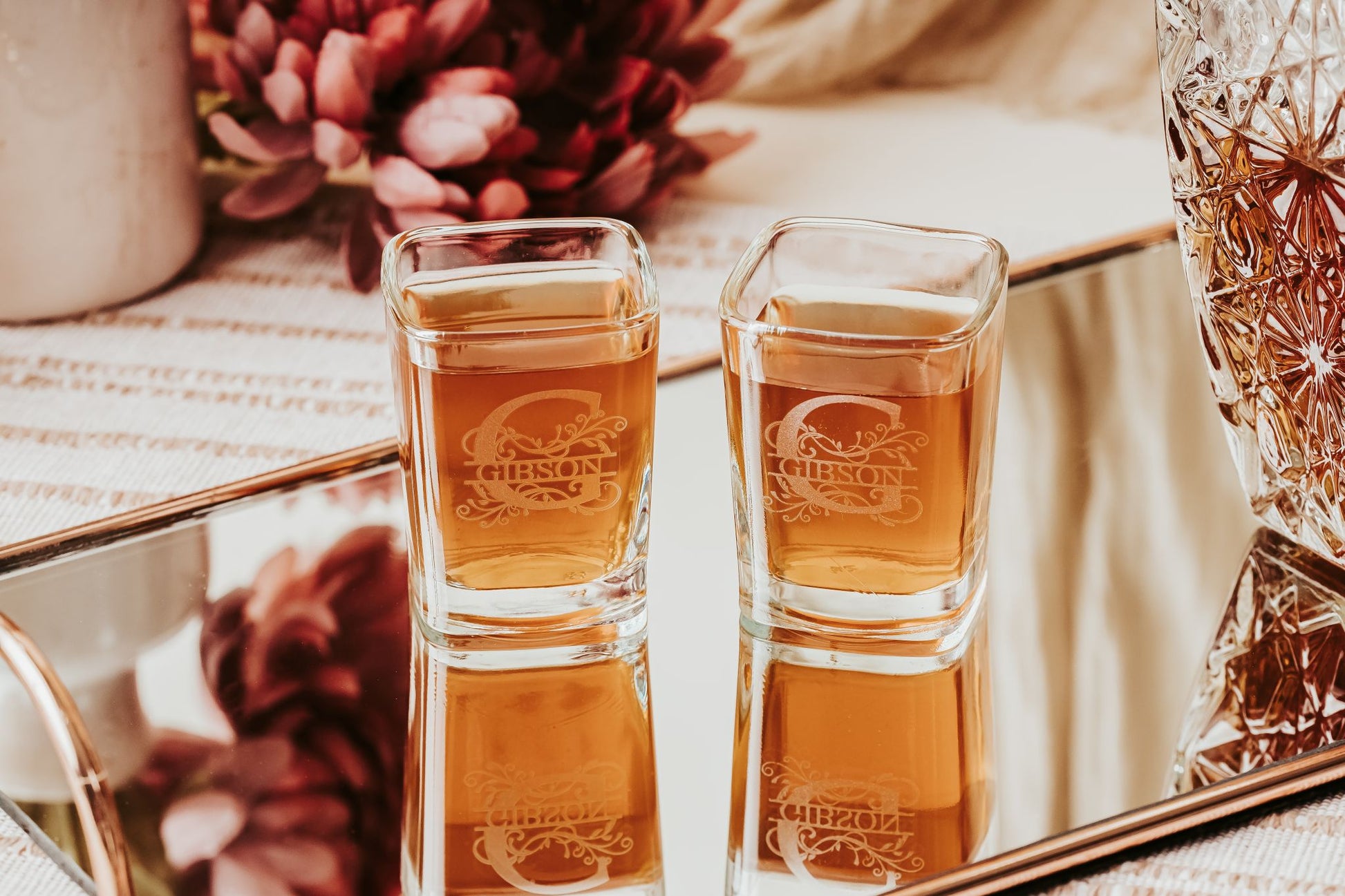 Two Personalized Monogram Shot Glasses by Mirabilia Boutique are filled with brown liquid and sit on a mirror, surrounded by pink flowers and a patterned decanter, creating an elegant gift set display.